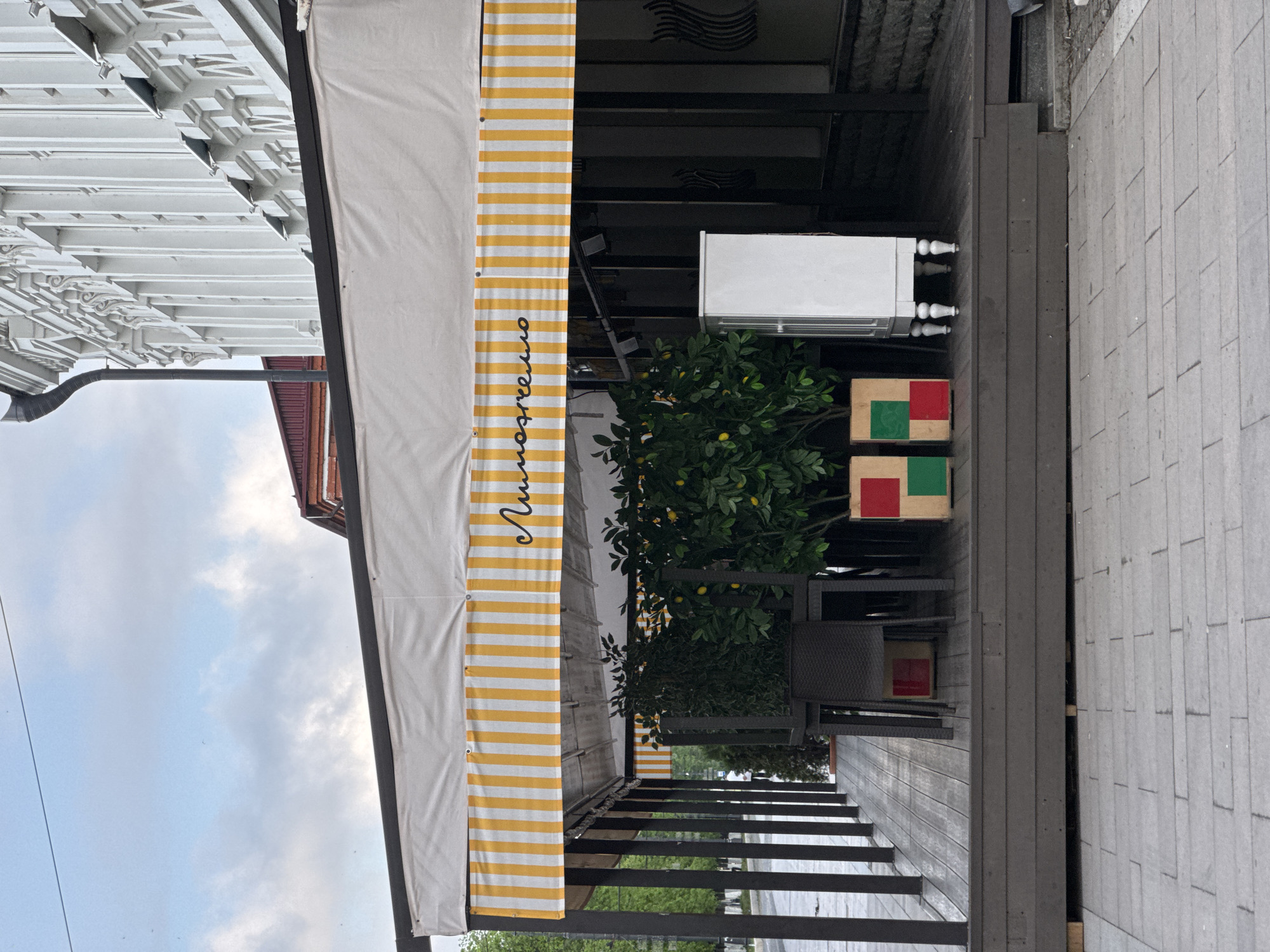 The image shows a cafe awning and outdoor seating area. The awning is white with yellow stripes and has the word "Липовечно" written on it. Below the awning is a plant with lemons. There are chairs and a small table with red and green squares on top. The seating area is on a wooden deck. The background shows a street with a sidewalk. The location is in Akhieli, Georgia.