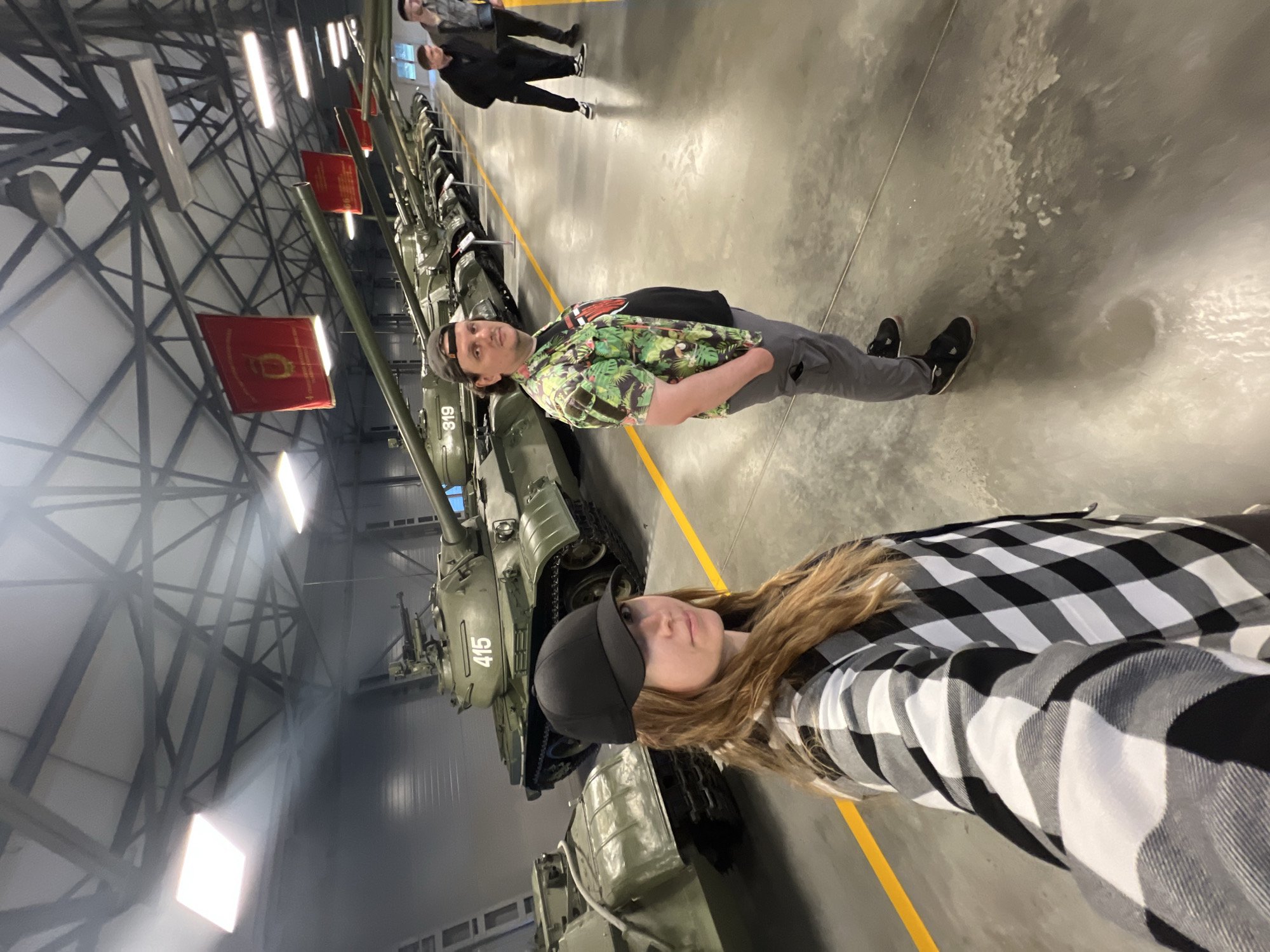 In an indoor exhibition hall, likely at the Patriot Park in Kubinka, Russia, two individuals are captured in a selfie. A woman in the foreground, wearing a black baseball cap and a black and white checkered shirt, looks towards the camera. Beside her, a man in a camouflage shirt and gray pants stands with his hands in his pockets. Behind them, a row of military tanks, painted in olive drab, are displayed, with one prominently featuring the number "415". The hall has a high ceiling with exposed industrial beams and red banners hanging overhead. The floor is concrete, marked with yellow lines. Several other individuals are visible in the background, observing the exhibits.