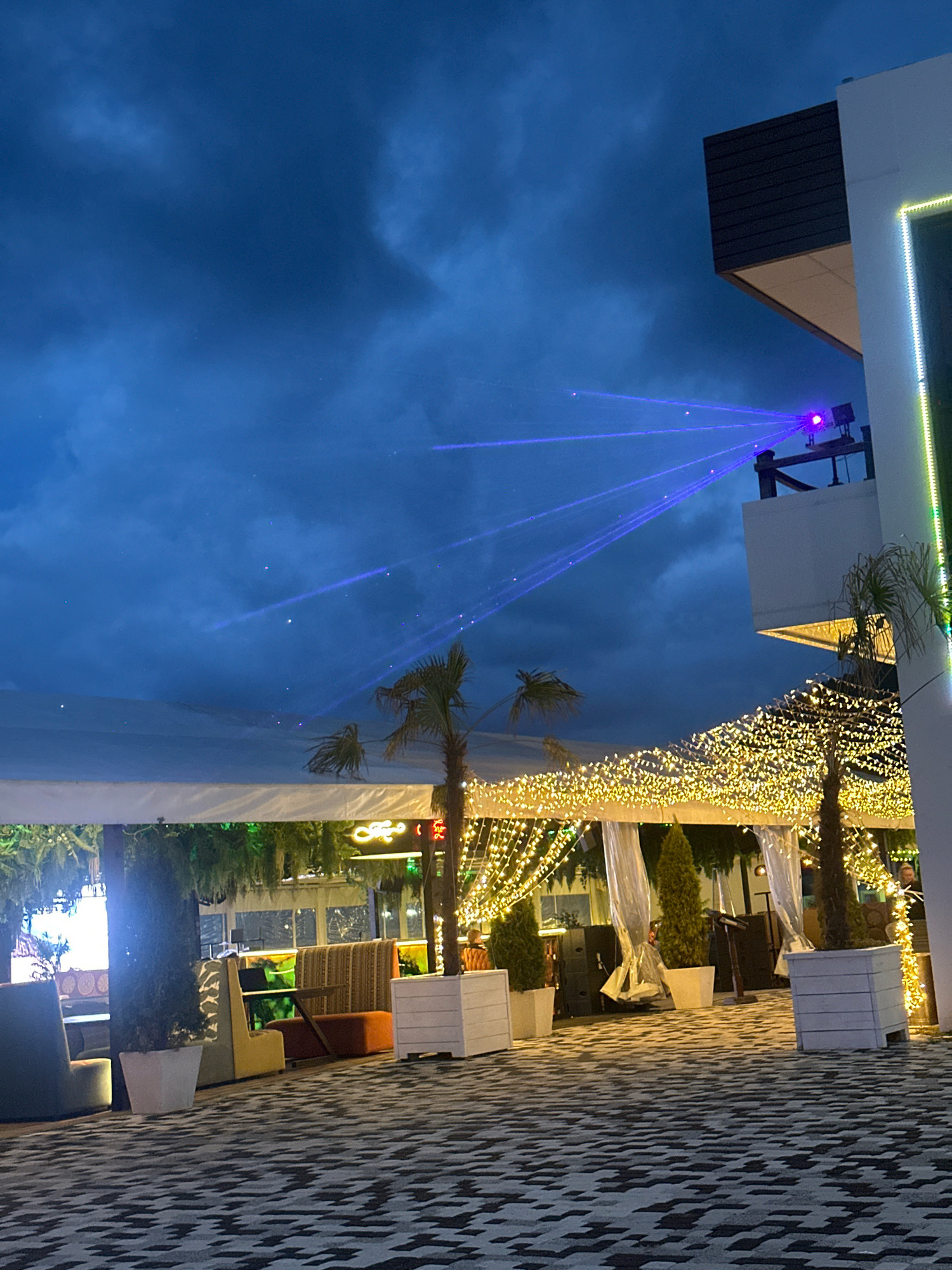 An outdoor establishment at night is depicted. A building with a modern design is visible on the right, featuring a balcony with a laser projector emitting blue beams into the dark, cloudy sky. The area below the balcony is decorated with numerous strings of yellow lights. Several palm trees and other plants are present, some in white planters. There are white canopies or tents, and seating areas with upholstered furniture and tables. The ground is paved with a geometric pattern. The scene appears to be a restaurant or entertainment venue.