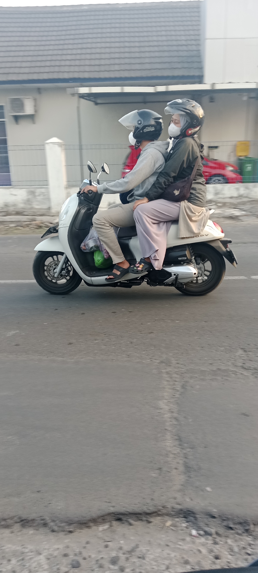 In Maguwoharjo, Indonesia, two individuals wearing helmets and face masks ride a white scooter on an asphalt road. The person in front, wearing a grey hoodie and tan pants, operates the scooter. The person behind them, wearing a dark jacket and light purple pants, has their arms around the driver. A black bag is slung across the passenger's body. A green plastic bag containing items is attached to the front of the scooter. In the background, a building with a gray tiled roof and an air conditioning unit is visible, along with a red car and a green trash bin. The scene is captured in daylight with a clear sky.