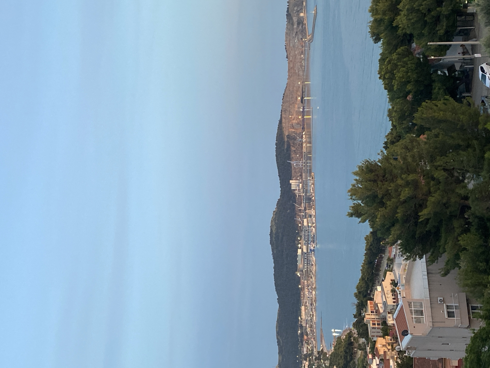 This is an elevated, eye-level, medium shot of a coastal city with a harbor at dusk. The sky is a pale blue with some faint cloud cover. The city is situated along a bay, with a line of buildings nestled between the water and a large, tree-covered hill in the background. The buildings are of various sizes and architectural styles, many with red-tiled roofs. A harbor with numerous boats and ships is visible in the middle ground, with cranes and industrial structures present on the far side of the bay, suggesting a port or industrial area. The water in the bay is calm, reflecting the sky and the lights of the city. In the foreground, several large evergreen trees obscure a portion of the view of the city. A few residential buildings are visible in the lower left quadrant, with balconies and windows. A white car is parked on a road in the lower right quadrant. The overall impression is of a peaceful evening scene in a coastal town.