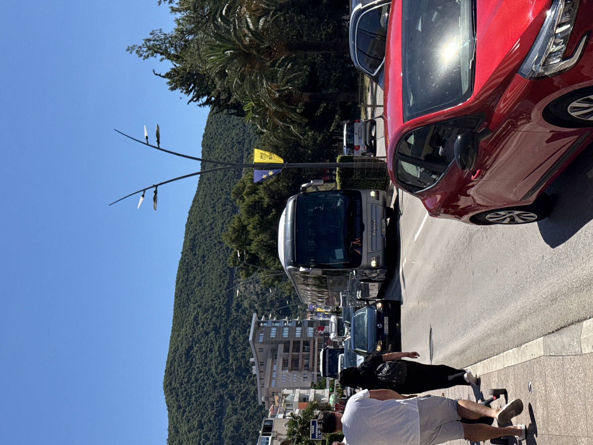 A traffic jam is depicted on a sunny day on a coastal road in Budva, Montenegro, with a view of a lush green mountain in the background. Several vehicles, including a large silver bus, a red car, and black cars, are stopped or moving slowly in the traffic. Pedestrians are walking on the sidewalk, some with luggage, suggesting a tourist area. Palm trees and other greenery line the street, and modern buildings are visible on the left side of the road. The clear blue sky indicates fair weather.