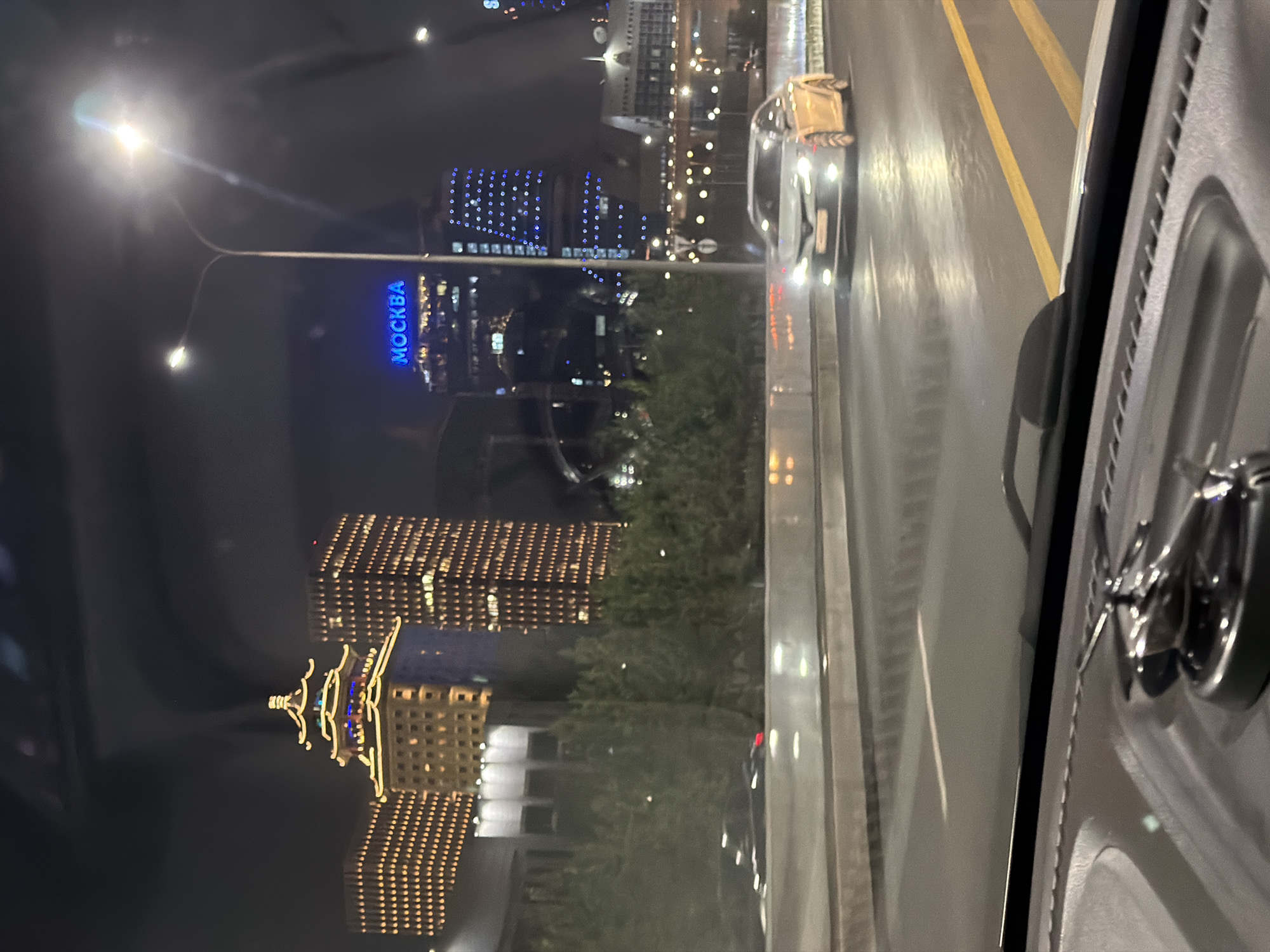 This image depicts a night view of a city skyline, likely Astana, Kazakhstan, as indicated by the text "MOCKBA" (Moscow in Russian) displayed on a building, which is a common feature in the city's architecture and often associated with its landmarks. The perspective is from inside a vehicle, looking out through the windshield. In the foreground, the dashboard of the car is visible, with a chrome ornament on top.
The scene is dominated by illuminated skyscrapers and buildings. One prominent building features a traditional East Asian-inspired roof design, also lit up with golden lights. Adjacent to it are taller, rectangular buildings covered in a grid of golden lights. Further to the right, another skyscraper displays a pattern of blue lights. A street lamp with a bright light source illuminates the upper right portion of the image.
The road in front of the car appears wet, reflecting the lights from the surrounding environment. A silver sedan is driving in the right lane, its headlights on. The road is marked with yellow lines. The background is dark, suggesting nighttime, and the sky is overcast or obscured by city lights. Green trees line the median or the side of the road in the middle ground. The overall impression is of a modern, vibrant city at night.