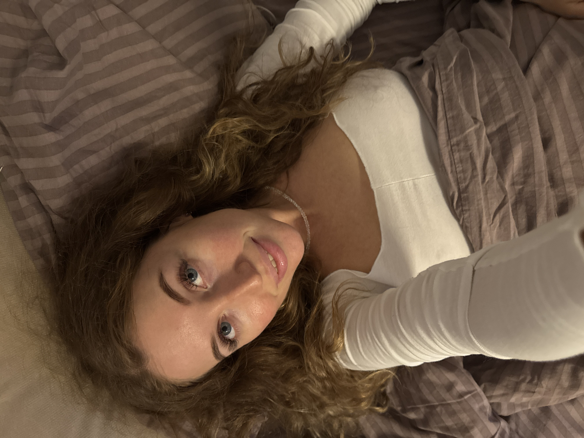A woman is lying on a bed. She has long, wavy brown hair, blue eyes, and is wearing a white long-sleeved shirt and a beaded necklace. She is looking up at the camera. The bed has striped bedding. The location is unknown.