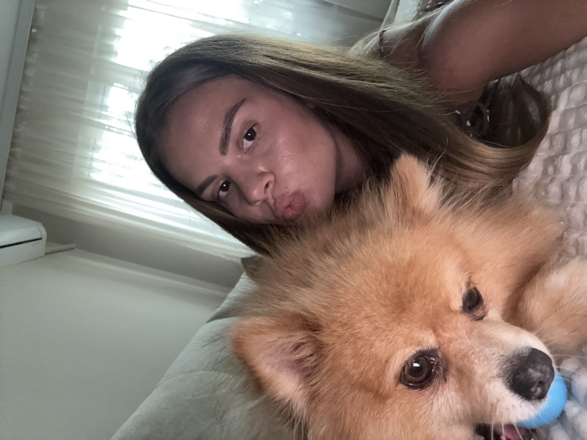 A Pomeranian dog and a young woman are lying on a bed. The dog is to the left, and the woman is to the right. The dog is facing left and has its mouth open, with a blue ball in its mouth. The woman is looking at the camera with her lips pursed. The background shows a window with sheer curtains and an air conditioning unit mounted on the wall. The scene appears to be indoors, in a domestic setting, likely a bedroom. No specific Plovdiv landmarks are visible.