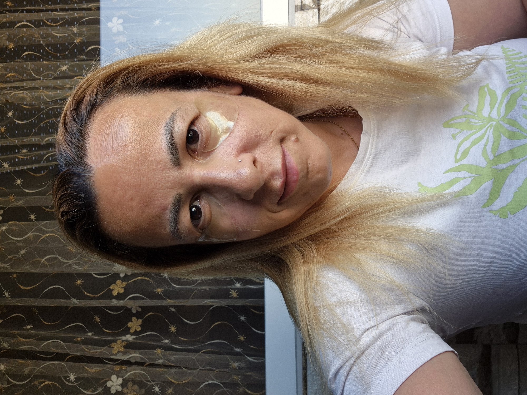 A close-up, medium shot shows a woman with blonde hair, wearing a white t-shirt with a green floral print. She has under-eye patches on and a small nose piercing. She is looking towards the camera with a slight smile. The background features a sheer, patterned curtain and a window with diffused light. The setting appears to be indoors, near a window, possibly in a residential area.