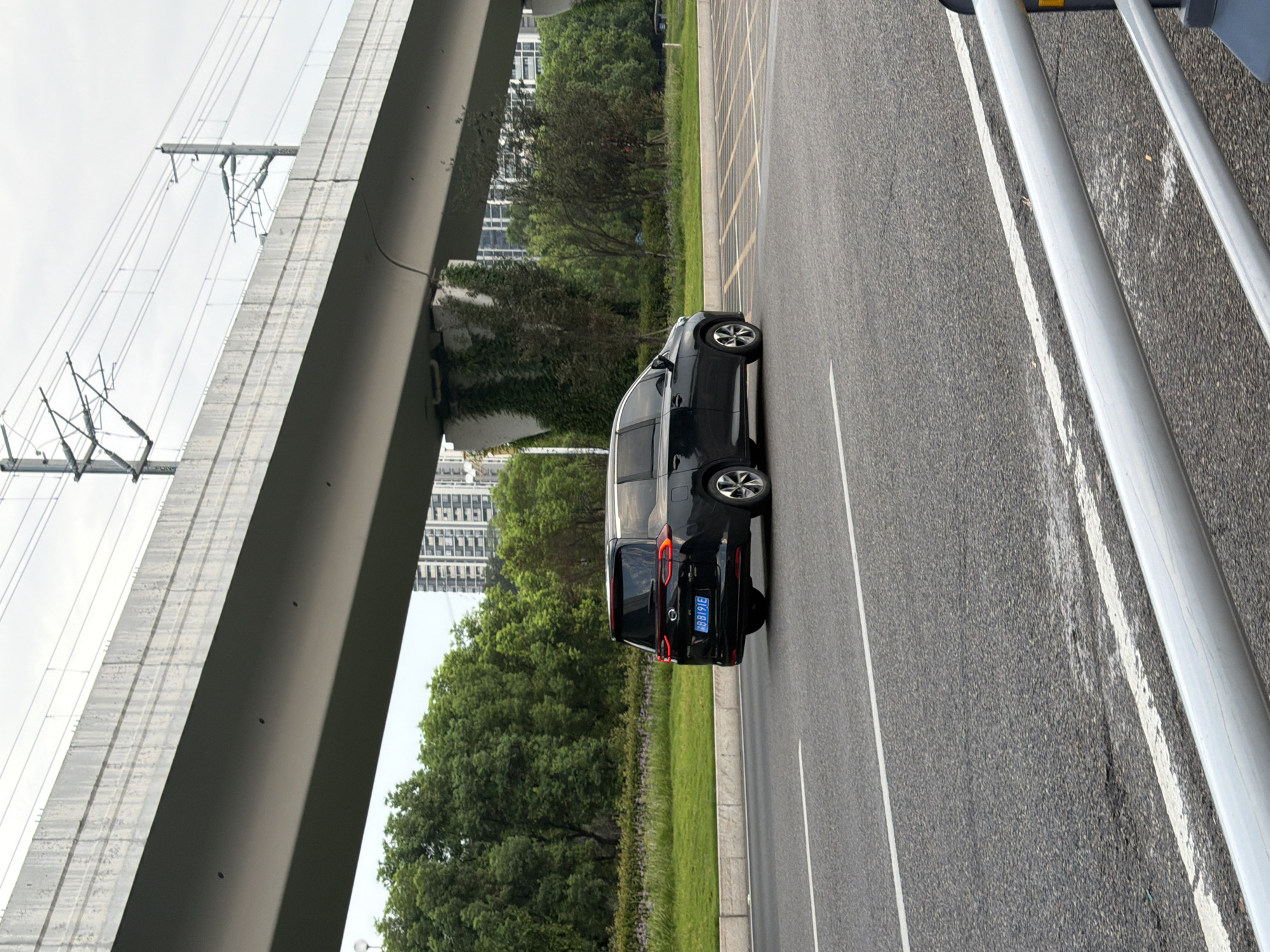 A black minivan with license plate "BB B191E" drives on a paved road under an elevated structure, likely a highway or railway overpass. The overpass is concrete, with visible support pillars covered in ivy. Overhead, power lines and their supporting poles are suspended from the overpass. Beyond the road, a grassy area with scattered purple flowers leads to a dense line of green trees. In the background, several multi-story buildings are visible. The road has white lane markings. The sky is overcast.