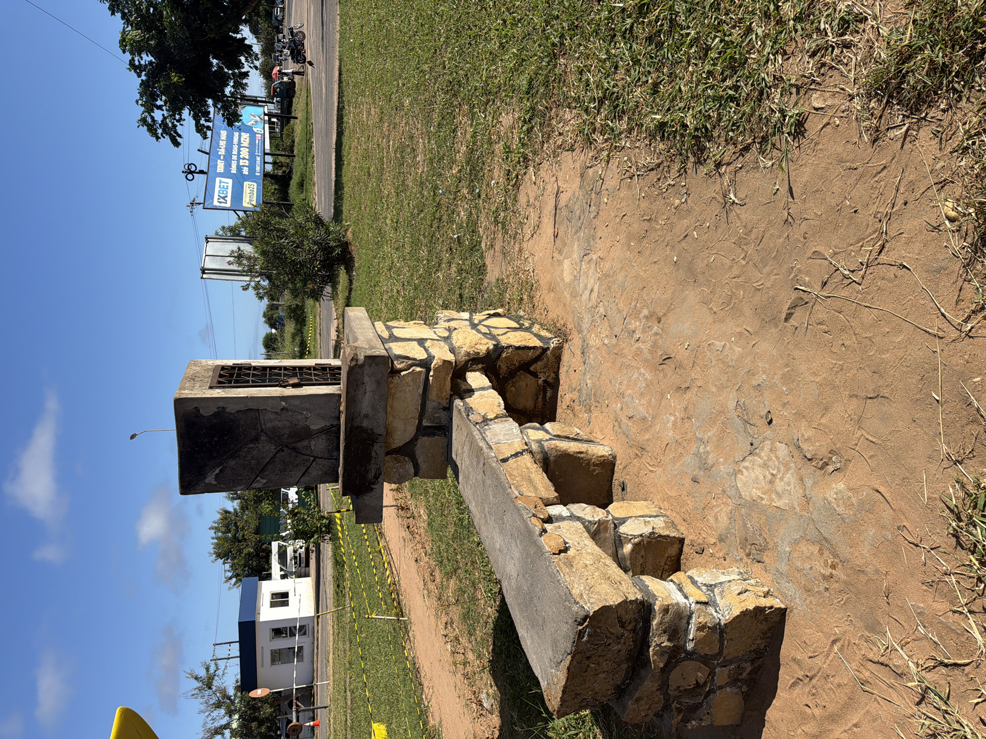 This outdoor image, rotated 90 degrees clockwise, captures a sunny daytime scene alongside what appears to be a road or street.
In the foreground, on the left side of the rotated image, a weathered, rustic stone and concrete structure is prominent. This structure is composed of irregularly shaped, light-colored stones cemented together, forming a base and what resembles a bench or low wall, with a taller, rectangular block at one end. The ground around the structure is a mix of reddish-brown dirt and patches of green grass. Faint tracks and patterns are visible in the dry dirt, suggesting pedestrian or small animal activity.
Extending towards the right side of the rotated frame, the ground transitions from dirt to a strip of vibrant green grass, bordering a paved road visible further in the background. Along the road, several vehicles, including motorcycles and cars, are visible, along with a few people. Trees and bushes line the roadside. In the middle-right of the image, a large blue billboard stands tall, partially obscured by tree foliage. Further into the background, on the far left, some buildings are discernible under a bright blue sky with scattered white clouds. A street light pole is also visible against the sky.
The weather is clear and sunny, indicative of midday or early afternoon. The visible text on the billboard provides a strong clue to the location: "1XBET - DÁ-LHE MAIS," "BÓNUS DE BOAS-VINDAS," "até 13 200 MZN!" (Mozambican Metical), "Pemba25," and "1XBET.COMZ." These details suggest the scene is in Mozambique, possibly near Pemba.