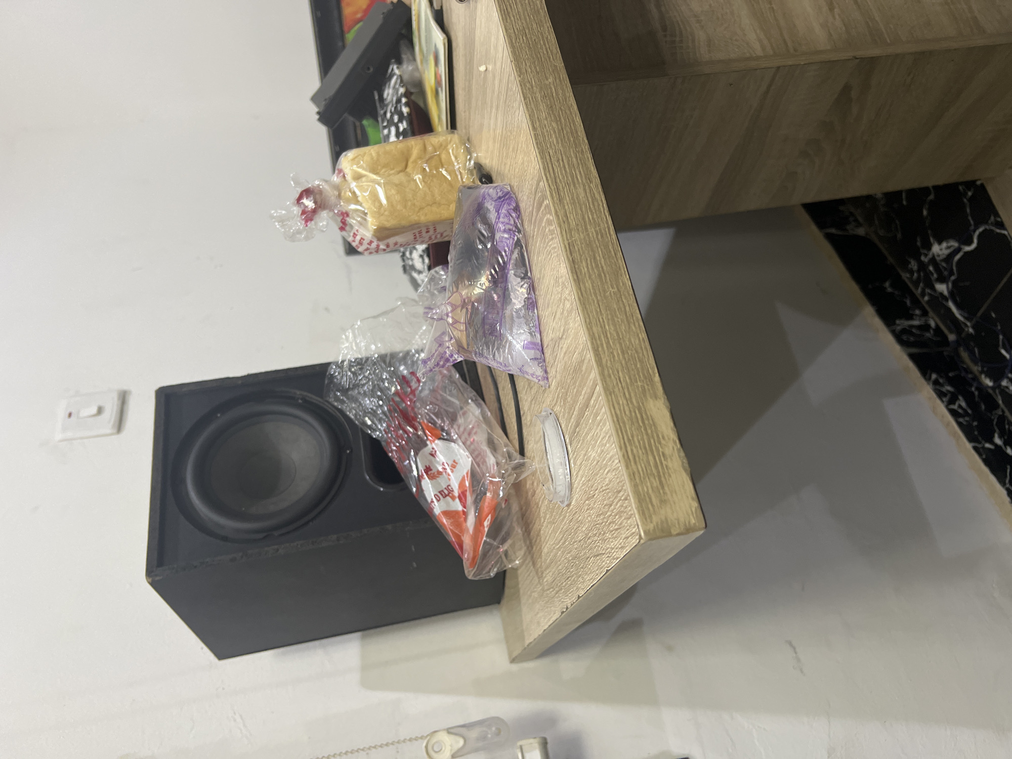 The image displays an indoor scene, taken at a tilted angle, showing objects arranged against a light-colored wall and on a wooden shelf or table. The wooden shelf, which would typically be horizontal, appears to extend diagonally upwards from the center-left towards the top-right due to the camera's orientation.
On or near the edge of this wooden shelf, several items are visible, mostly contained within clear plastic bags. These include a small, golden-brown loaf of bread in a clear bag with red and white printed details, and another item encased in a distinctive purple clear plastic bag. Further down, another clear plastic bag holds a red and white cone-shaped object, on which faint, partially obscured text, possibly "OULIG," is visible.
Below and to the left of the wooden shelf, a black rectangular speaker or subwoofer with a prominent circular speaker cone is placed against the wall. A white electrical light switch is visible on the wall further to the left. A small, clear, circular adhesive hook is attached to the underside of the wooden shelf. In the background, beneath the shelf, a surface with a black and white marble pattern can be seen, possibly part of another piece of furniture or flooring.
The setting appears to be a room, potentially in Abuja, Nigeria, as provided in the context. There are no people present, and no particular activity is taking place; the image captures a static arrangement of items. The time of day and weather are not discernible.