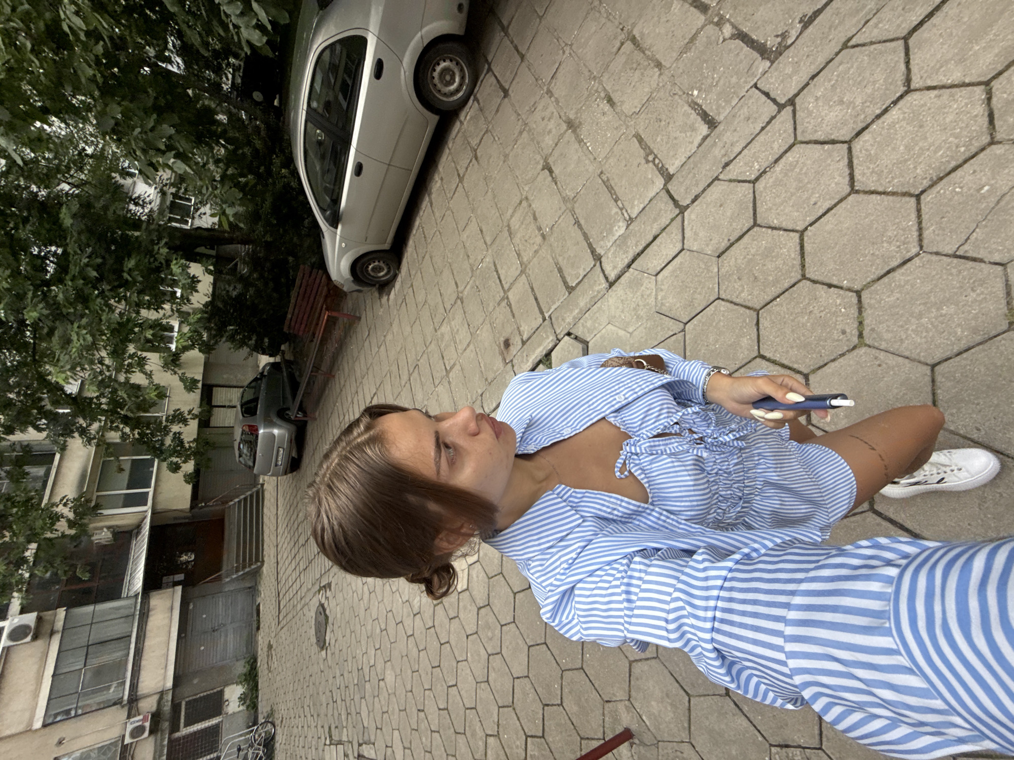 A young woman with brown hair styled in two buns is standing on a hexagonal paved area in front of a multi-story apartment building. She is wearing a blue and white striped short-sleeved shirt over a matching bustier and shorts. She is holding a white electronic cigarette in her right hand and looking to her left. To her left, there is a bicycle parked near the entrance of the building. The building has multiple windows and doors, and there are trees with green leaves visible. A silver car is parked to the right of the woman, and a red bench is situated near it. The overall setting appears to be a residential courtyard in the city of Plovdiv, Bulgaria, given the architectural style of the building and the surrounding urban environment.