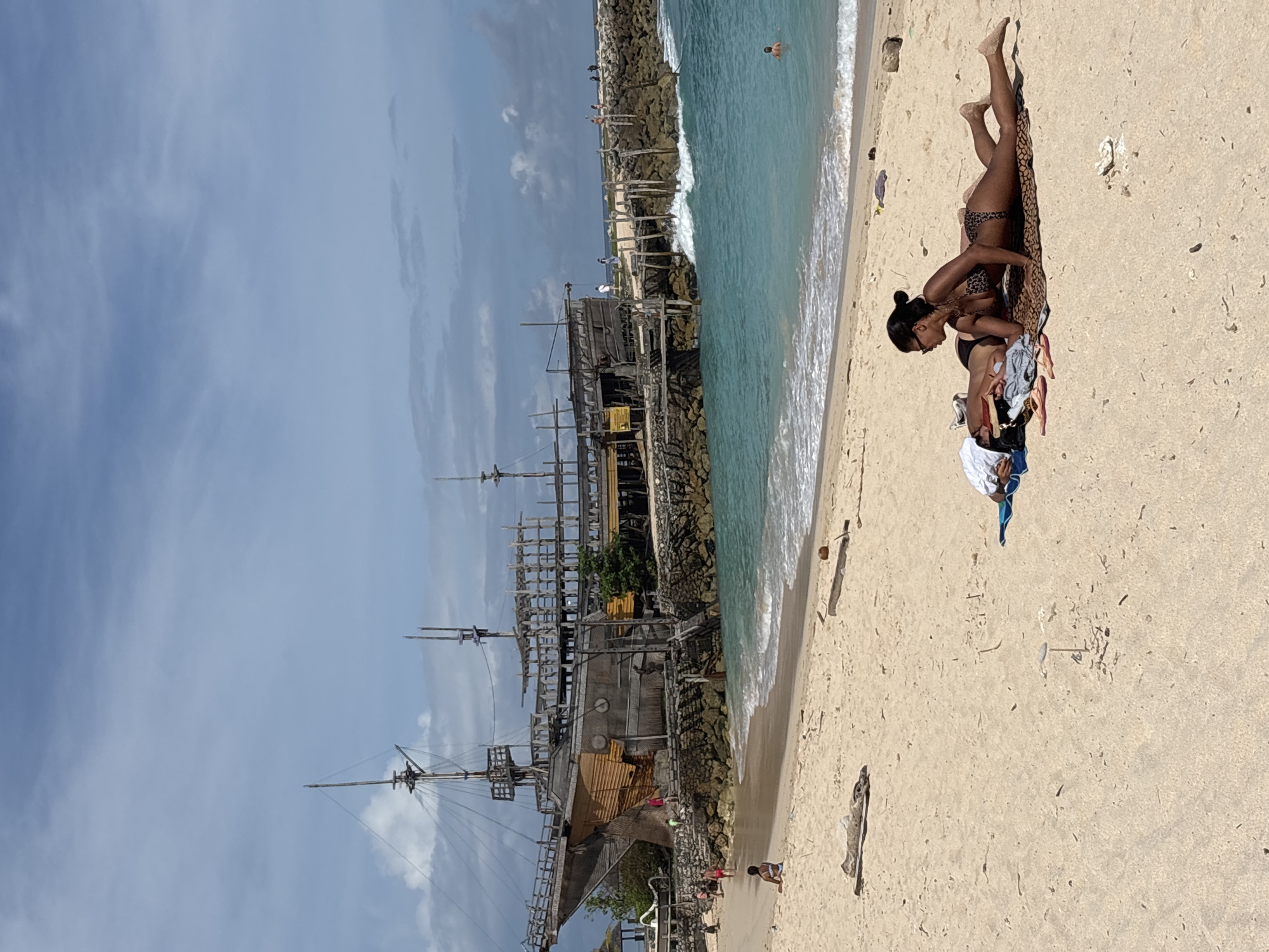 The image shows a sunny beach scene in Kuta Selatan, Indonesia, next to the Pirate Bay Bali restaurant. A woman in a leopard-print bikini lies on a towel on the sand near the water's edge. Some clothing is visible near the woman. The restaurant, a large wooden structure resembling a pirate ship, stands inland from the beach. Waves are visible in the water. People are on the beach and swimming in the ocean.