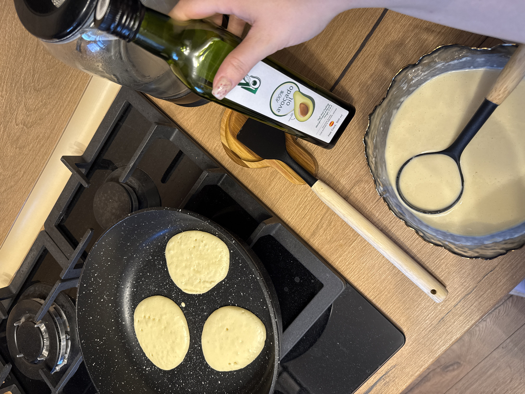 A top-down view shows a person preparing pancakes on a gas stovetop. Three pancakes are cooking in a black speckled frying pan on the left side of the frame. To the right of the stovetop, a hand is holding a green glass bottle of "100% avocado oil" over a wooden spoon rest that contains a black spatula. A bowl filled with pancake batter and a ladle is to the right of the spoon rest. The stovetop is black and stainless steel, and the countertop is a light wood grain.