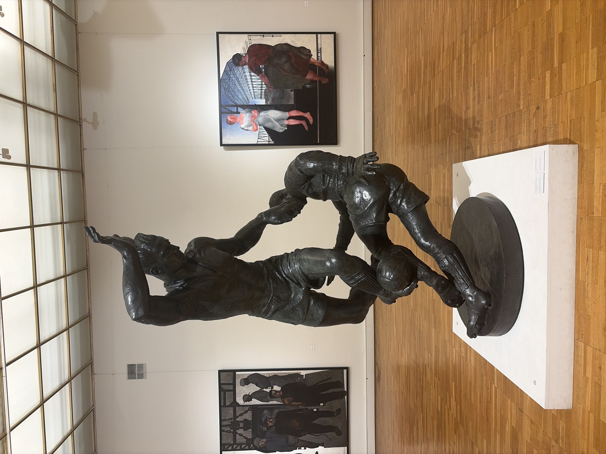 This media file displays a photograph taken inside an art gallery, likely in Moscow, Russia. The primary subject is a large, dark bronze sculpture depicting two men engaged in a dynamic, athletic pose, seemingly playing a sport such as football or rugby. One figure is in the foreground, lunging forward with a ball at his feet, while the other is behind him, reaching out with one arm. Both figures are muscular and appear to be wearing athletic shorts. The sculpture is mounted on a white rectangular pedestal, which itself sits on a polished wooden floor.
Behind the sculpture, two framed artworks hang on a plain white wall. The upper artwork is a painting featuring two figures in a dimly lit setting with industrial or architectural elements in the background. The lower artwork is also a painting, depicting a group of men in darker attire, again with a stark, possibly industrial background.
To the left of the sculpture and paintings, a section of a large window or skylight structure is visible, characterized by a grid of square panes with brass-colored framing. The lighting in the gallery appears to be ambient, coming from the ceiling and possibly the large window structure, suggesting it is daytime. The overall atmosphere of the scene is one of serious artistic presentation, with the focus on the sculptures and paintings displayed. There is no visible text in the image that provides specific information about the artworks or the gallery.
