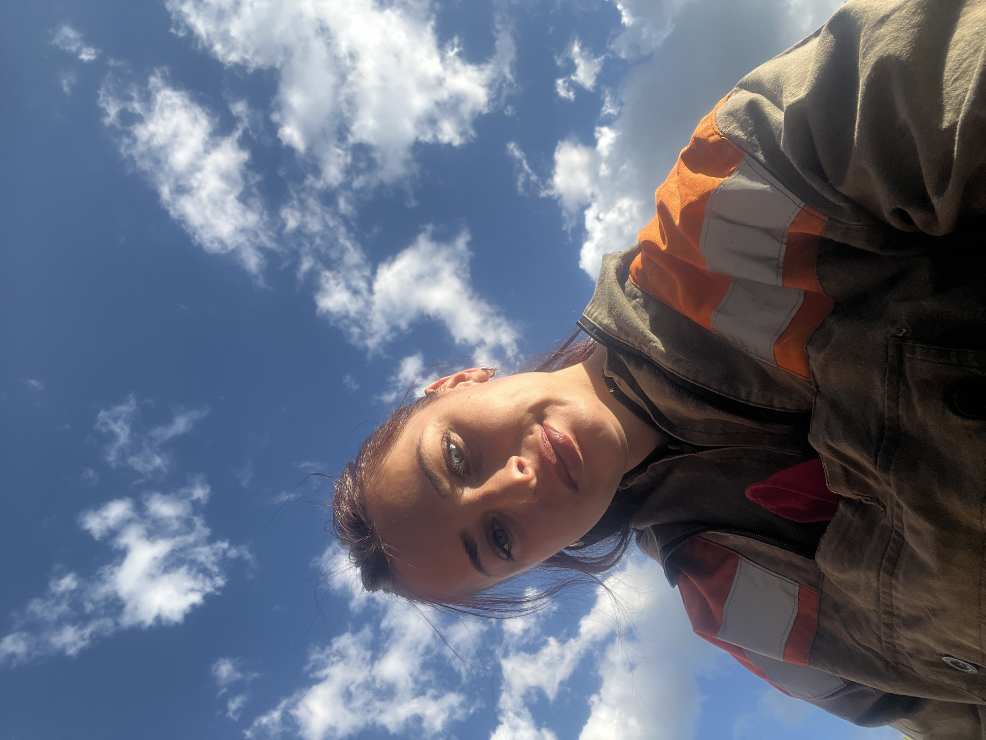 A low-angle portrait shot of a woman with auburn hair and light eyes, wearing a brown and orange jacket. The backdrop is a bright blue sky with scattered white clouds. There is no contextual information in the image to determine if the location is Chaikovsky, Russia.