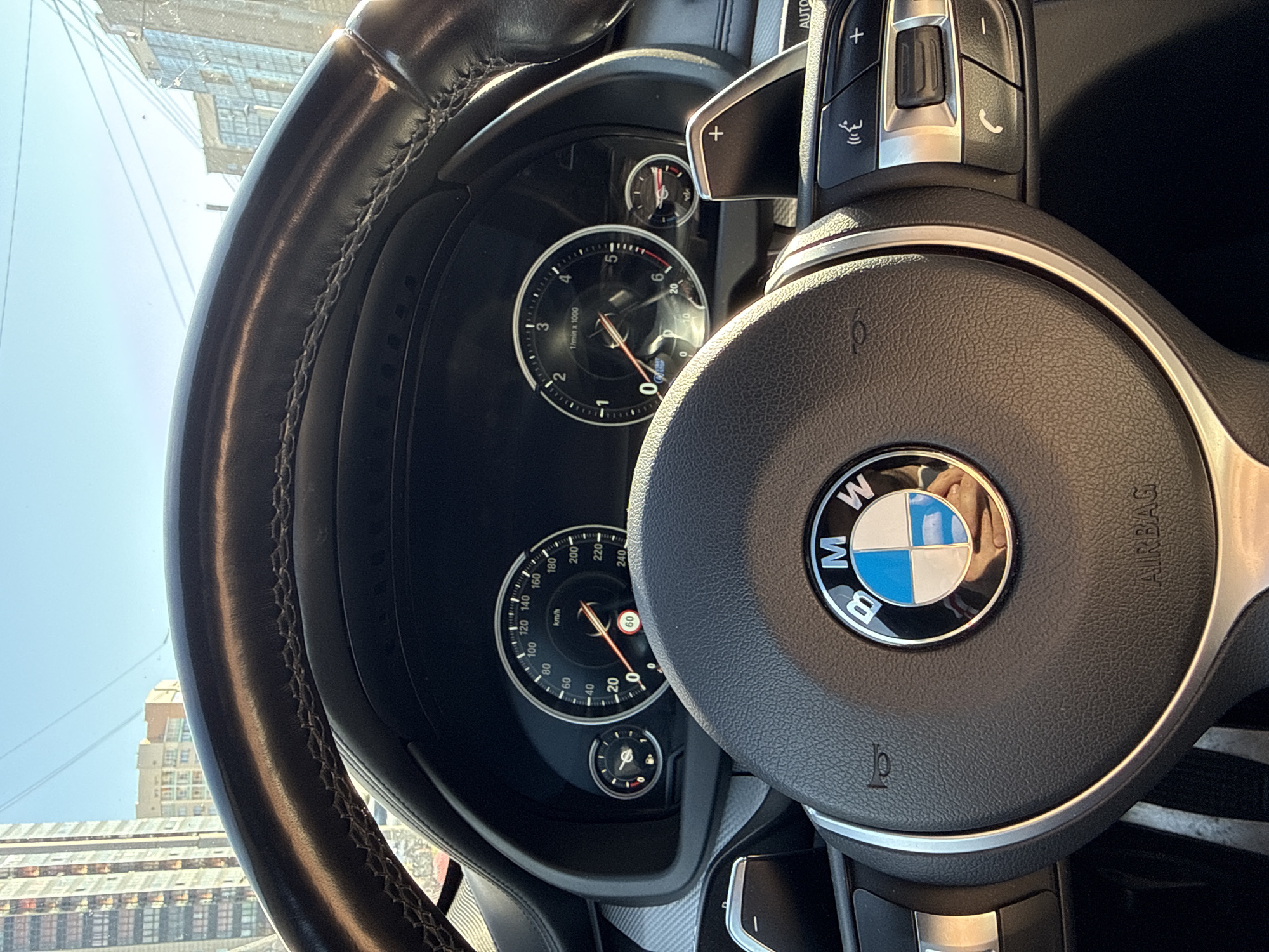 The image depicts a BMW steering wheel and dashboard. The BMW logo is prominently displayed in the center of the steering wheel. The dashboard features gauges displaying speed, revolutions per minute (RPM), and other vehicle information. The speedometer indicates a speed of approximately 55 km/h, while the tachometer reads close to 0 RPM. Outside the vehicle, buildings are visible. These buildings, when visually cross-referenced, align with structures in Krasnoyarsk, Russia, suggesting the photograph was taken in that city. There is a reflection of a person's hands in the BMW logo.