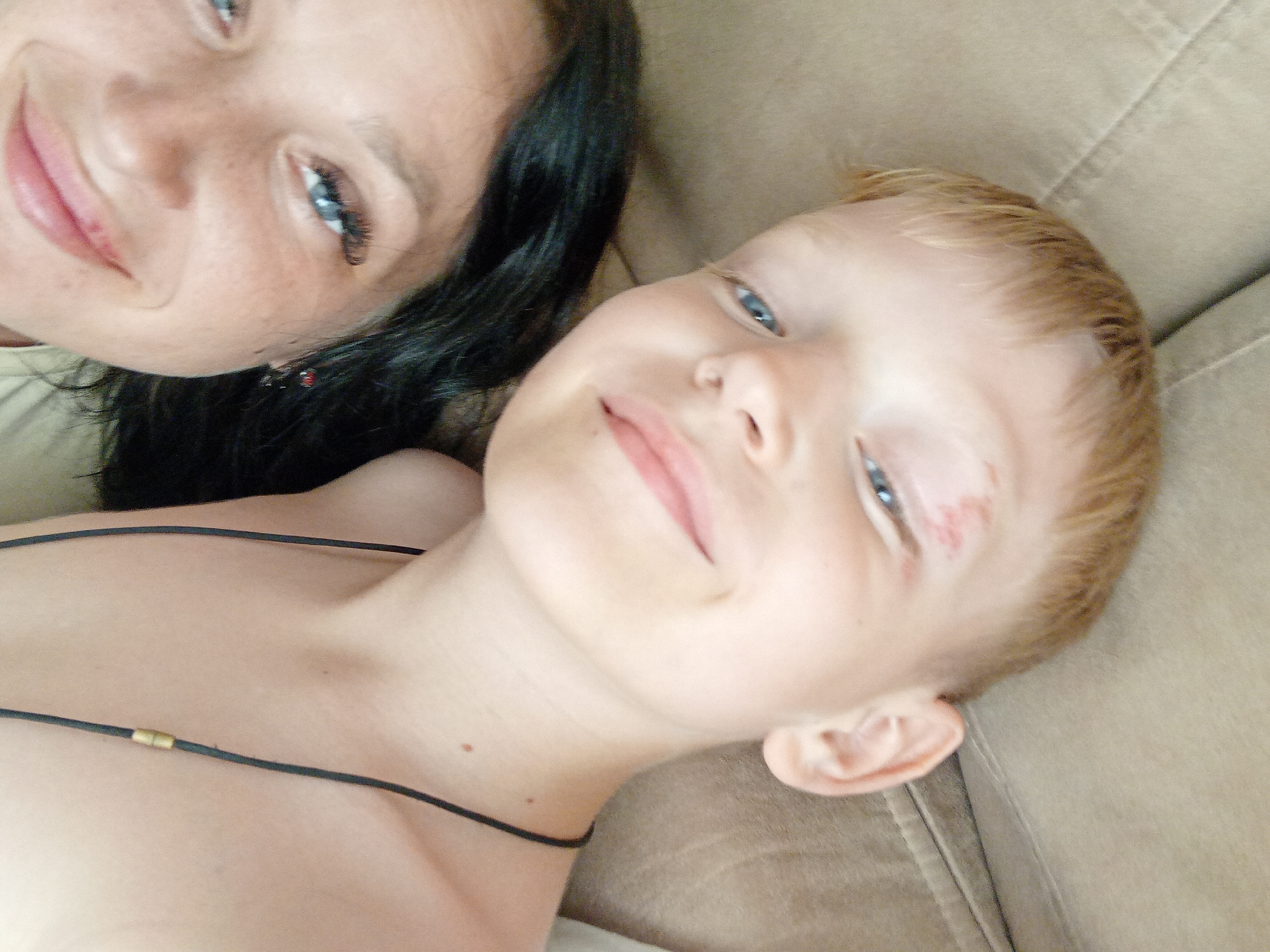 A close-up, high-angle shot shows two individuals, an adult and a child, lying down on a tan, tufted surface, possibly a sofa or bed. The adult, a woman with dark hair and blue eyes, is on the left, her face tilted towards the camera. She has fair skin with some freckles and is wearing a light-colored shirt. The child, a boy with blonde hair and blue eyes, is on the right, also looking towards the camera. He has a small red mark or scrape on his forehead. The child is wearing a black cord necklace with a small gold clasp. The background consists of the tan surface, which appears to be fabric with stitching. The overall lighting is soft and natural. The image is taken in Chuhuiv, Ukraine.