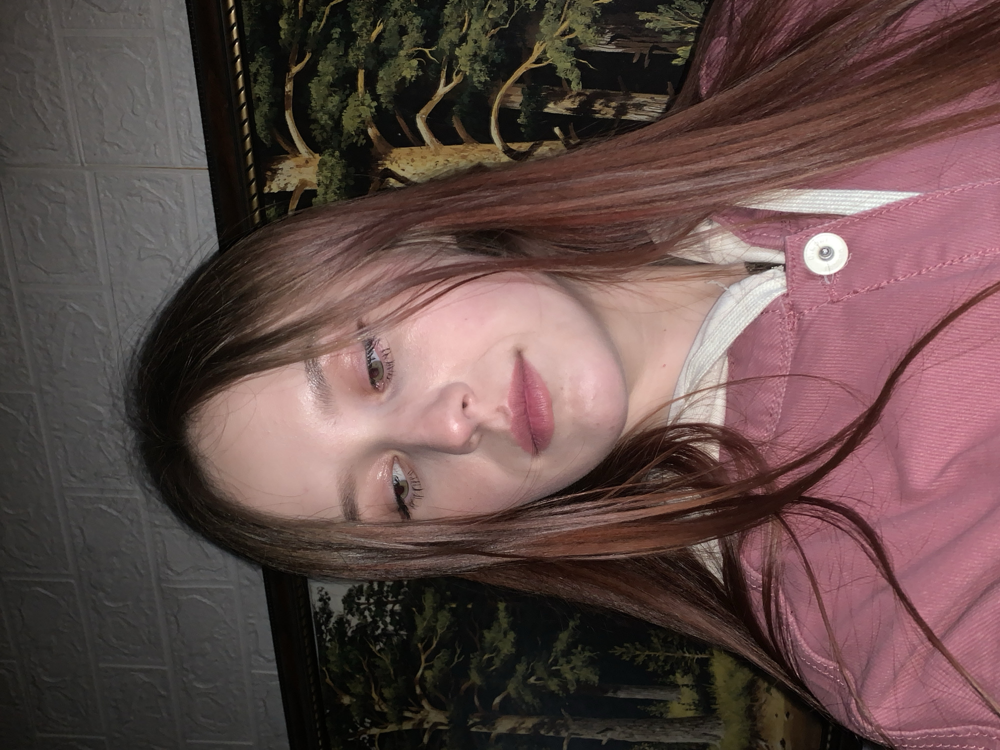 A woman with long, reddish-brown hair is the primary subject of this portrait-style image. She is wearing a pink denim jacket with a white collar visible underneath. Her face is slightly angled towards the camera, and she has light skin, dark eyebrows, and light-colored eyes.
The background features two distinct textures: a textured, white wall on the left and a framed painting or print on the right. The painting depicts a landscape scene with trees.
There is no information in the image to suggest that the location is in the city of Фоки, Russia.