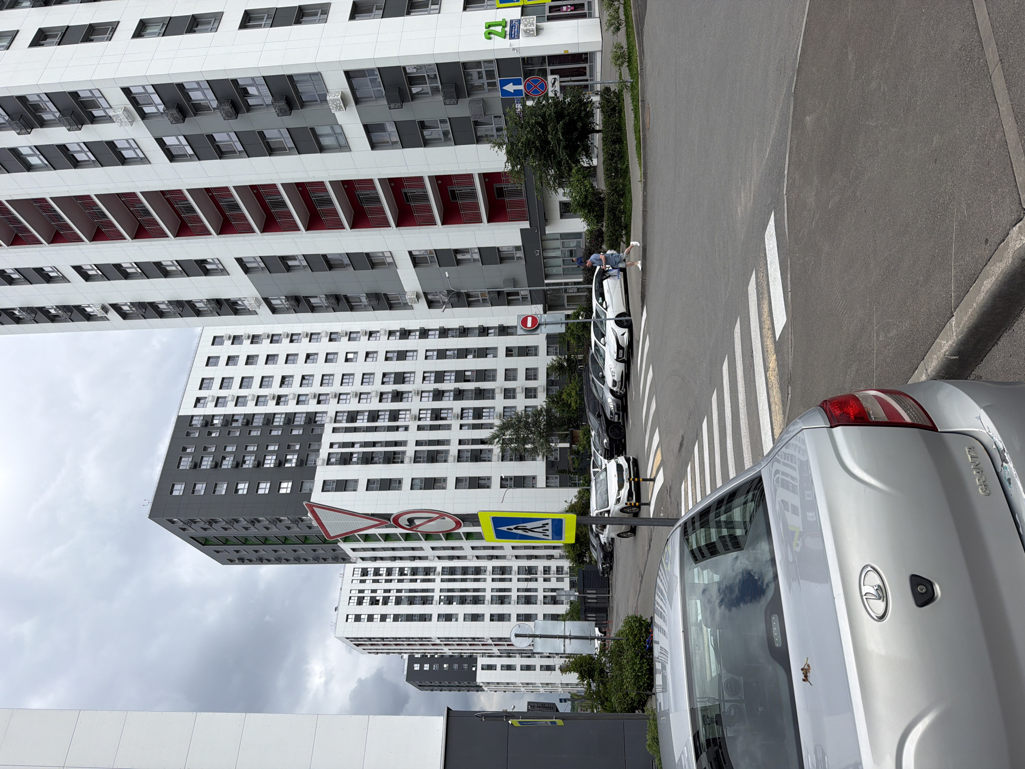 The image shows a street scene with modern apartment buildings and cars. A person in a blue shirt and shorts is standing next to a white car. Several other cars are parked along the street. There are traffic signs visible, including a pedestrian crossing sign and a "no entry" sign. The buildings are tall and multi-story, with white and grey facades. The sky is overcast, suggesting cloudy weather. The sun is not visible, but there is ambient light, indicating it is daytime. The overall impression is an urban environment. Some text is visible on the signs, but it is not clearly legible.