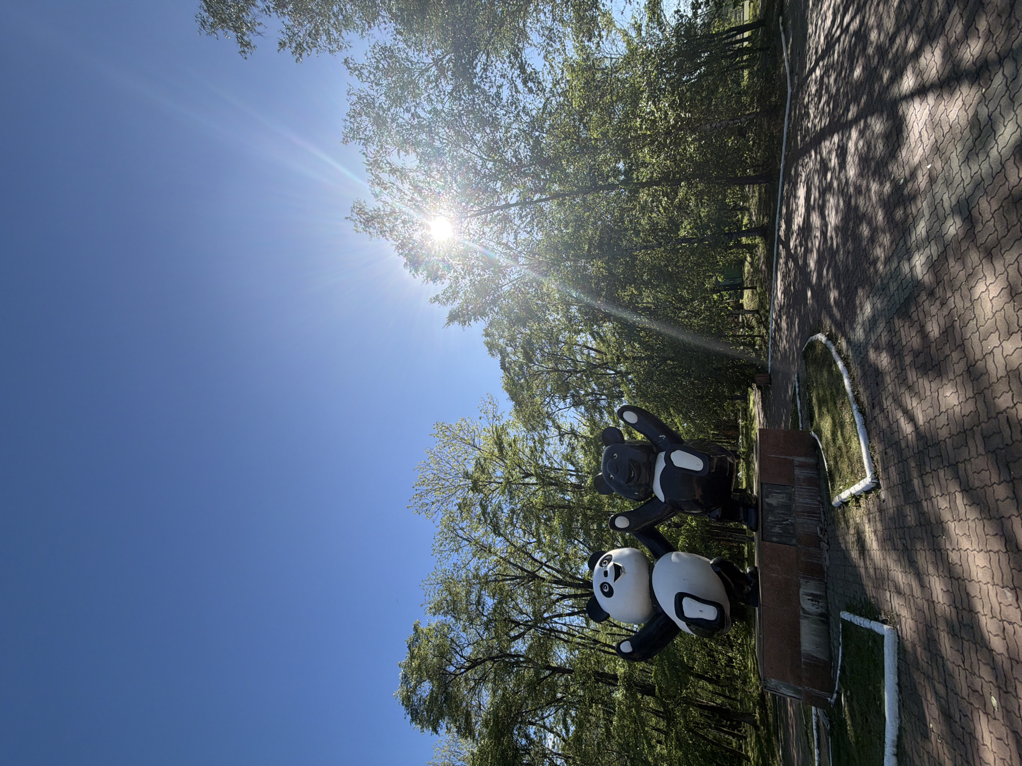 The image shows a sunny outdoor scene with two panda bear statues on a brick pedestal in a park setting. The park is located in Birobidzhan, Russia. The lower panda is white with black markings around its eyes and on its limbs, while the upper panda is primarily black with white markings. The sky is a clear blue with sunlight filtering through the trees in the background. A paved pathway is visible next to the statue base.