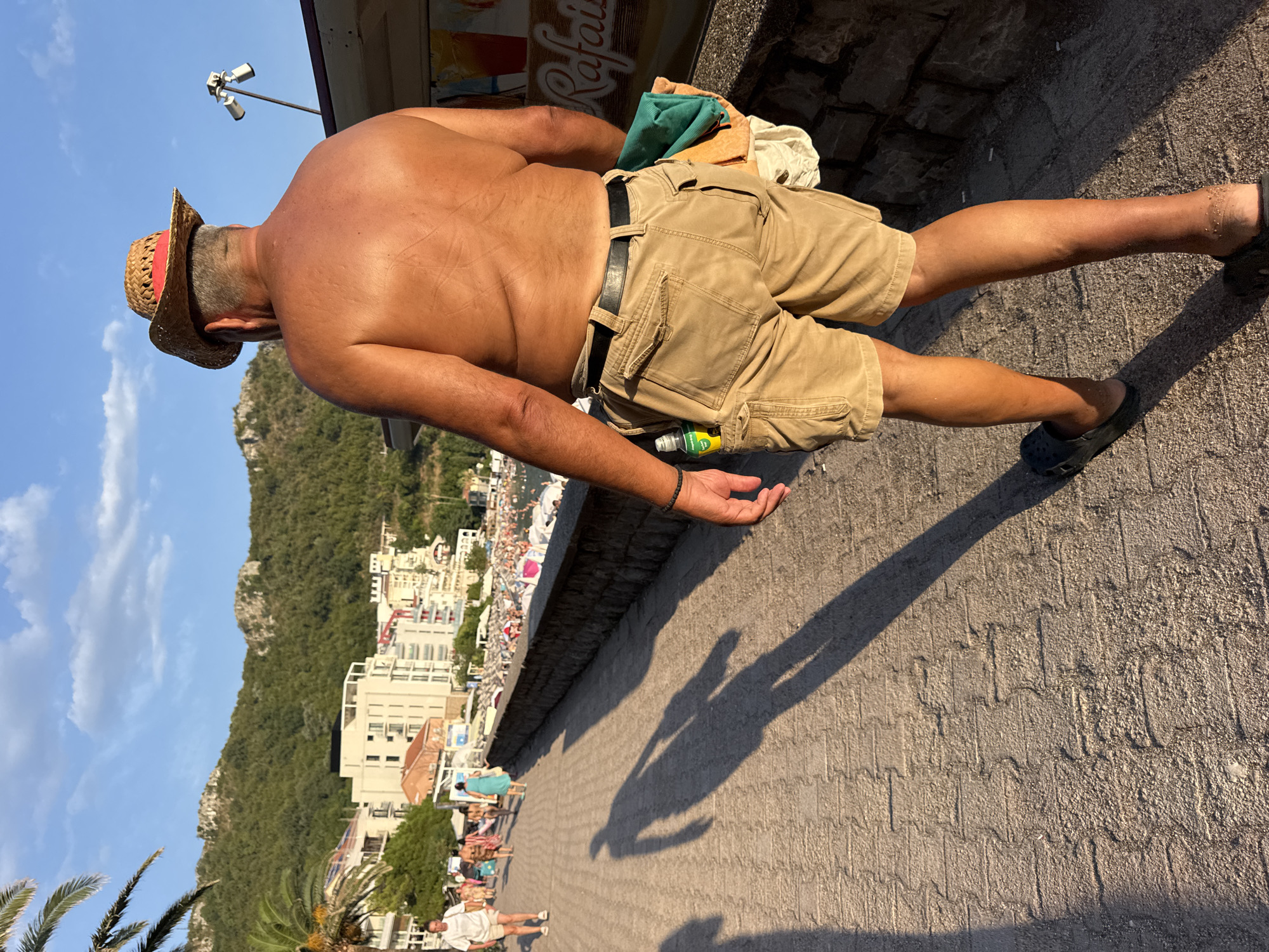 A man wearing a straw hat and tan cargo shorts walks away from the camera along a paved walkway in Rafailovići, Montenegro. The man is shirtless, revealing a tanned back. He has a black belt and black clogs on his feet. A green towel and other items are tucked into his shorts. In the background, there are buildings, a beach filled with umbrellas and people, and green, hilly terrain under a bright blue sky with some clouds. The sun is casting a long shadow of the man on the walkway.