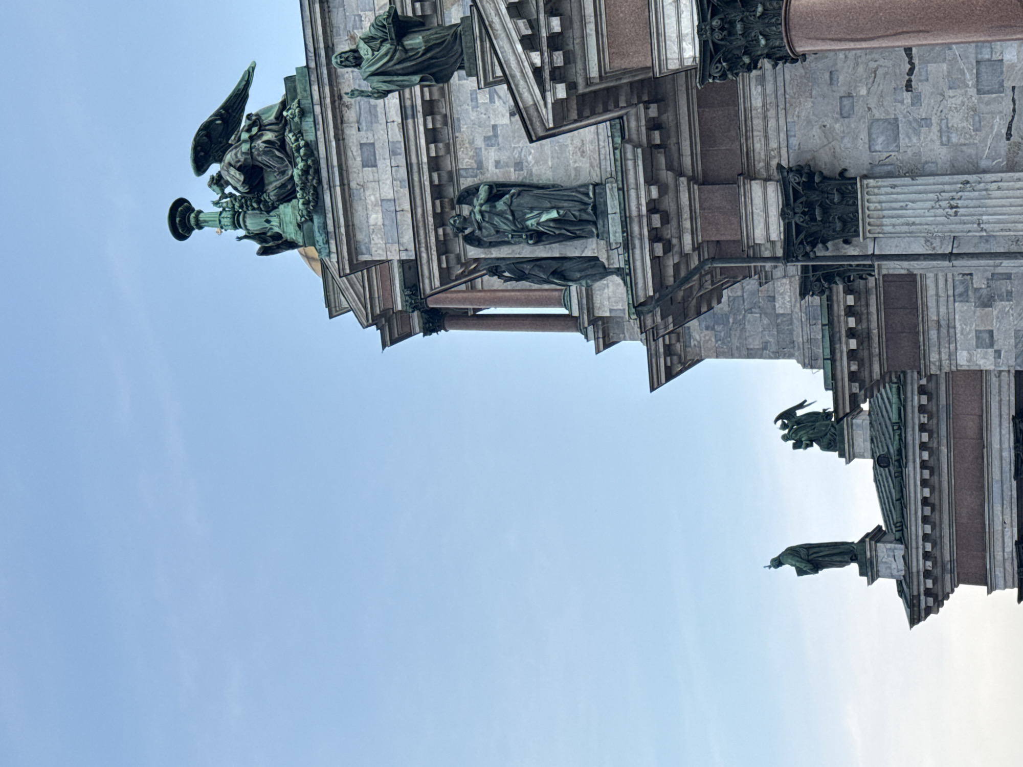 This is a low-angle shot of the exterior of Saint Isaac's Cathedral in Saint Petersburg, Russia, focusing on its architectural details and statuary. The sky is a clear, pale blue. The cathedral's facade is constructed of large, pale gray marble blocks.
Prominently featured are several large, green-patinated bronze statues. At the very top right, a statue of Jesus Christ stands with his right hand raised in a gesture of blessing and his left hand holding an open book. Below and to the left of this, on a higher ledge, is a statue of an angel with large wings, kneeling beside a decorative urn or lamp. To the left of the angel, on a lower ledge, stands another statue of an angel with outstretched wings, holding a cross. Further down and to the left, on the roof of a lower section of the building, are two more statues: one of a figure holding a cross and book, and another of two figures, one of whom has wings.
The building's architectural elements include Corinthian columns with intricate bronze capitals, cornices, and pediments. The roofs of the lower sections are covered with green metal. The overall impression is one of grandeur and religious significance.