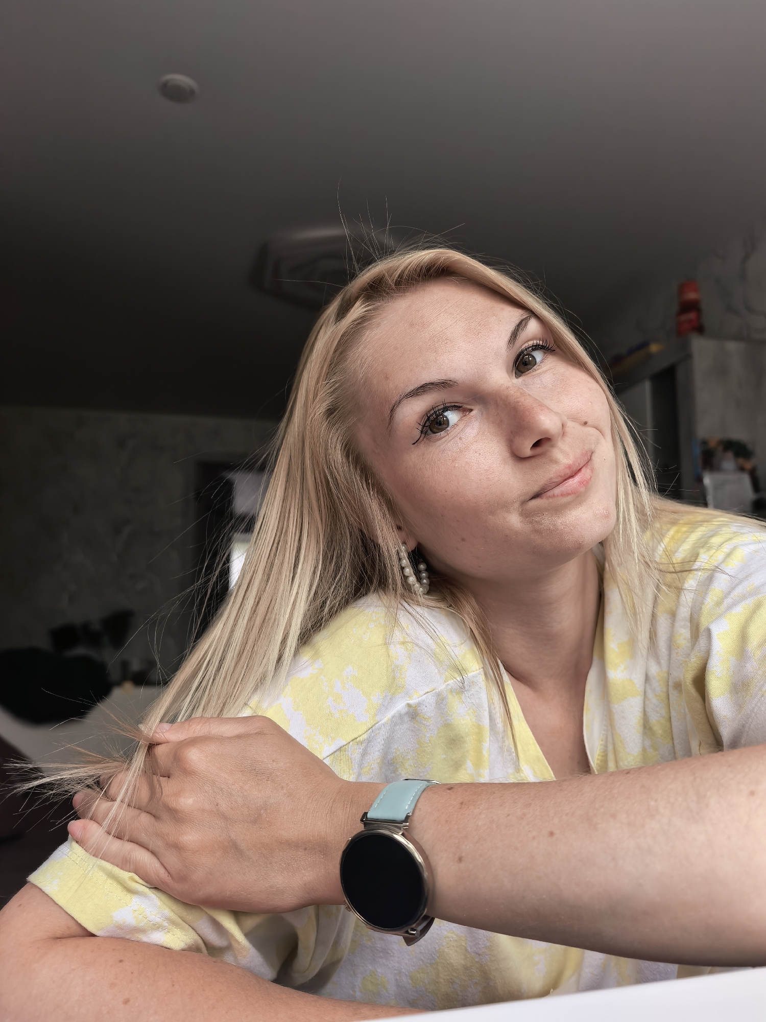 A blonde woman with freckles is facing the camera. She is wearing a yellow and white shirt and a watch with a light blue band. Her hair is long and straight, and she is holding a few strands of it in her hand. She appears to be indoors. The location in the city of Sterlitamak, Russia is not discernible from the image.