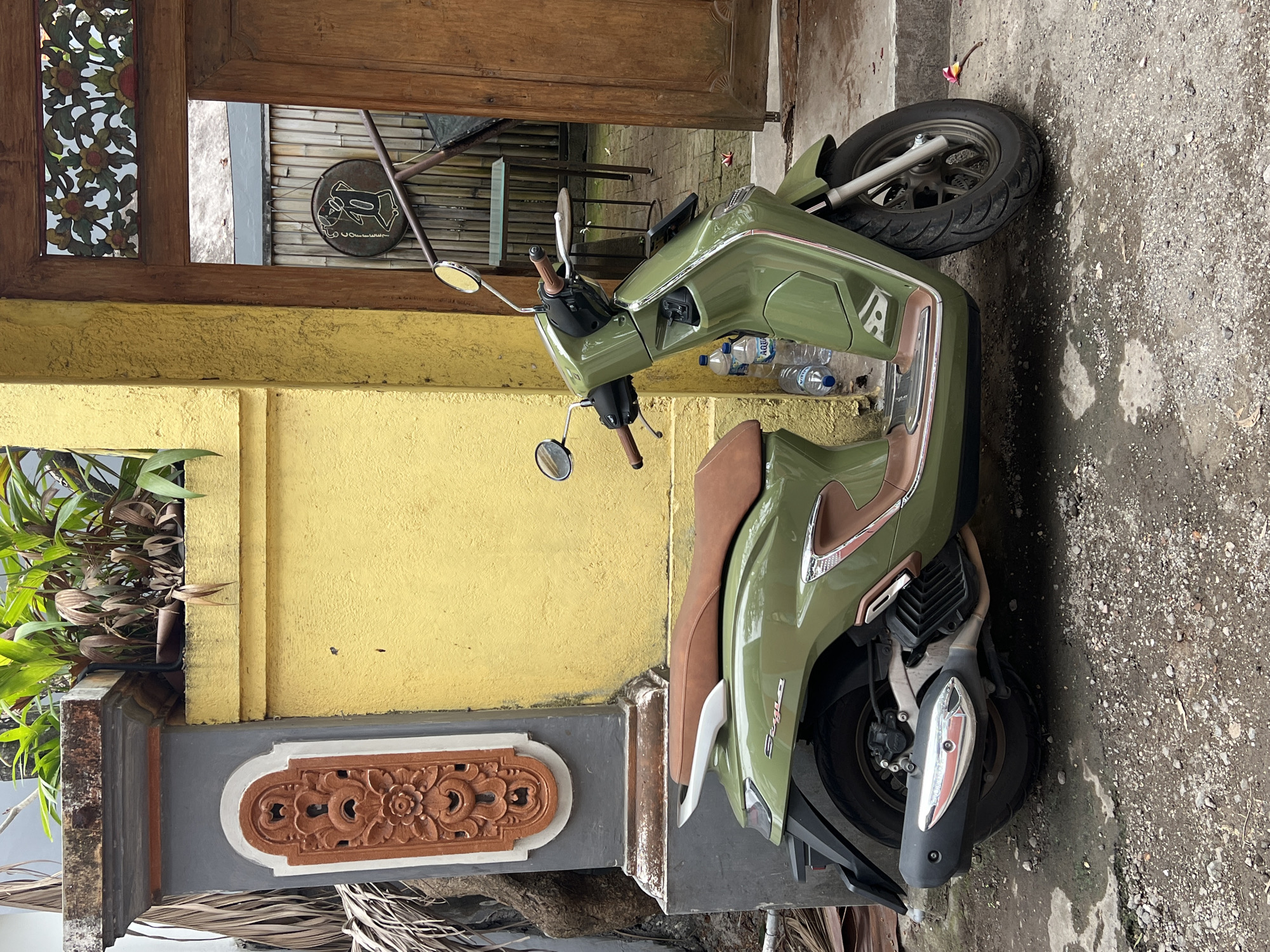 An olive-green scooter with a brown seat is parked on a gravelly and wet surface in front of a yellow wall and a weathered, wooden door in Denpasar, Indonesia. The scooter is a modern model with chrome accents. Two plastic bottles of "AQUA" brand water are on the ground near the scooter. To the left of the scooter is a grey pillar with decorative orange carving and a lush green plant on top. The wooden door is open, revealing a glimpse of an interior with bamboo-like fencing and a round sign with a figure. The ground is a mixture of dirt, small stones, and puddles, indicating recent rain. A single pink flower lies on the ground near the front wheel of the scooter. The overall impression is one of a quiet, slightly humid urban environment with a touch of tropical charm.
