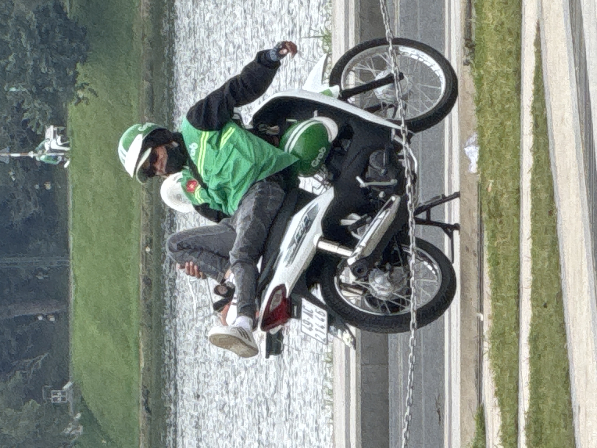 A person wearing a green Grab uniform, helmet, and sunglasses sits on a white motorcycle. The person's legs are extended forward, with one foot resting on the motorcycle's handlebar. The motorcycle is parked on a paved surface next to a body of water, likely Xuan Huong Lake in Da Lat, Vietnam. The background shows a grassy slope with trees and a few distant figures. The scene appears to be in a public park or recreational area.