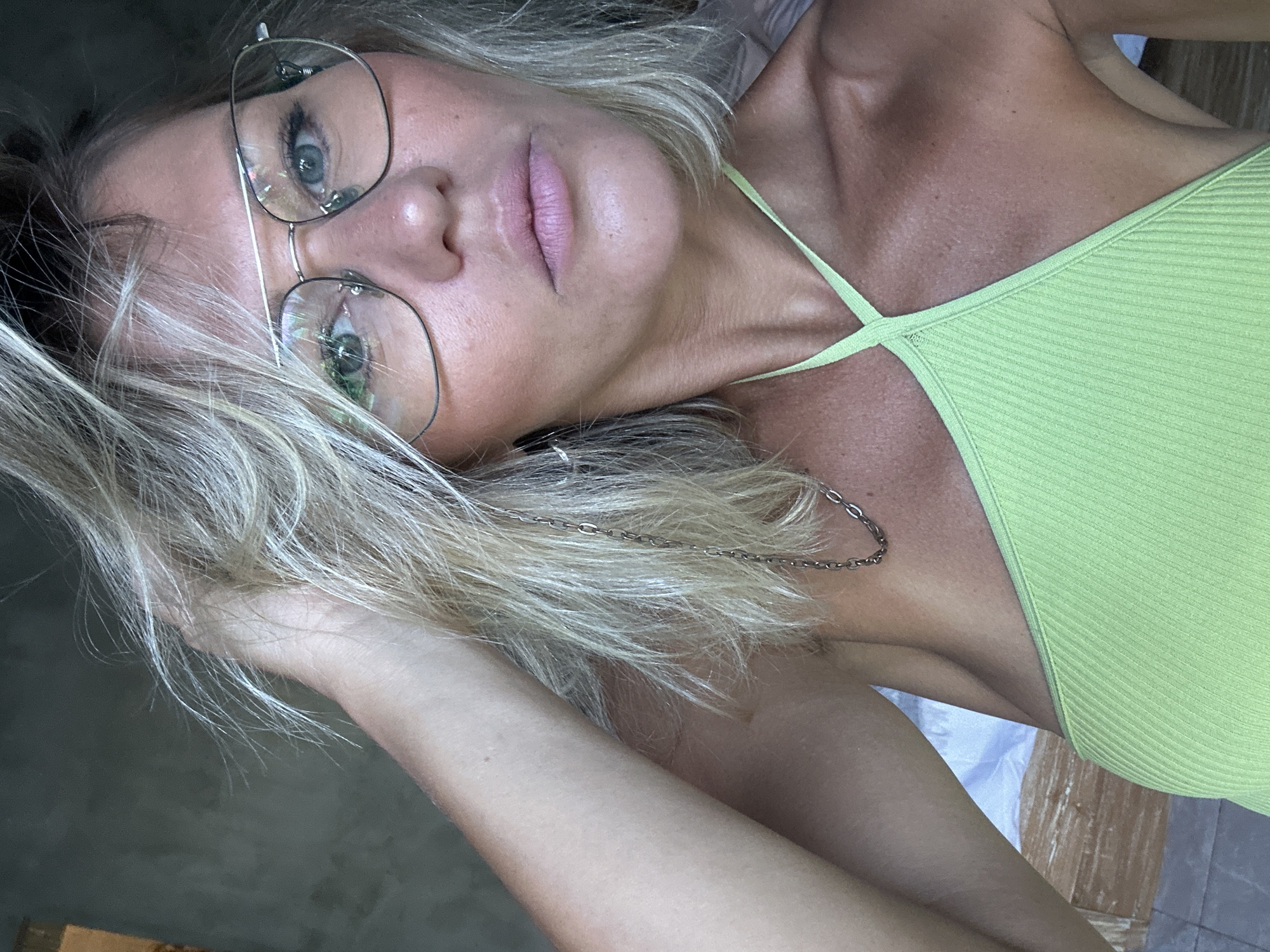 A fair-skinned woman with blonde hair and glasses is shown from the chest up, looking directly into the camera. She is wearing a lime green halter top and a silver chain necklace. Her left hand is raised, touching her hair. The background is out of focus, but appears to be an indoor setting with a bed and a wall. The lighting is bright, casting shadows on her face and neck. This image was taken in North Kuta, Indonesia.