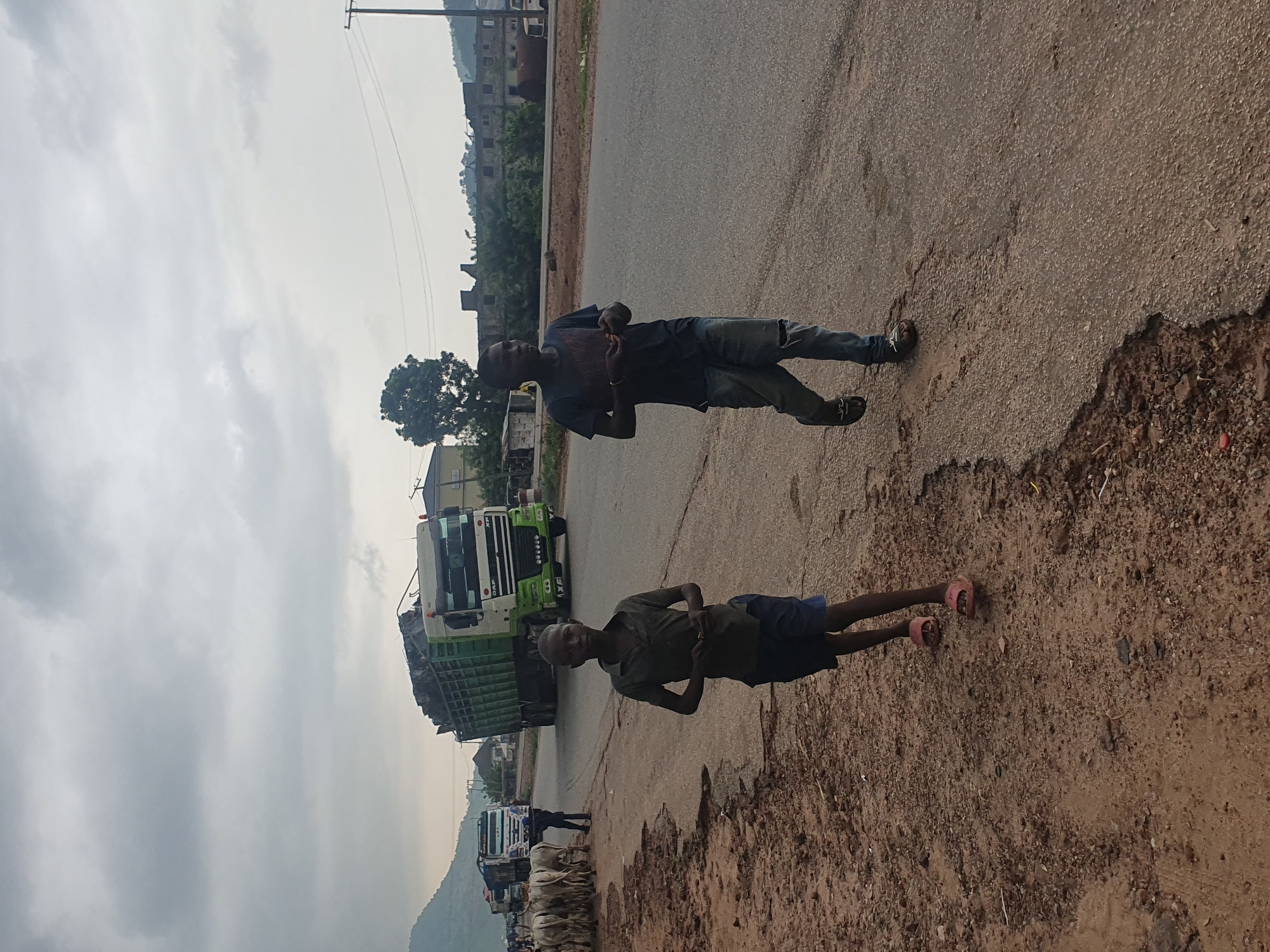 Two young boys walk along a dusty roadside in Lokoja, Nigeria. The boy on the left wears a dark green t-shirt and blue shorts, with pink sandals. The boy on the right is dressed in a dark blue t-shirt and ripped blue jeans, also wearing sandals. They appear to be walking in the same direction. In the background, a large green and white truck is parked on the side of the road, with its cargo bed filled with what appears to be scrap metal or debris. Several other trucks are visible further down the road, along with a herd of cows standing near the side of the road, some with a person tending to them. Buildings and trees are scattered along the horizon, with a hazy sky overhead, suggesting an overcast day. The road itself is paved but shows signs of wear and tear, with potholes and cracks. The ground beside the road is a mixture of dirt, gravel, and sparse vegetation.