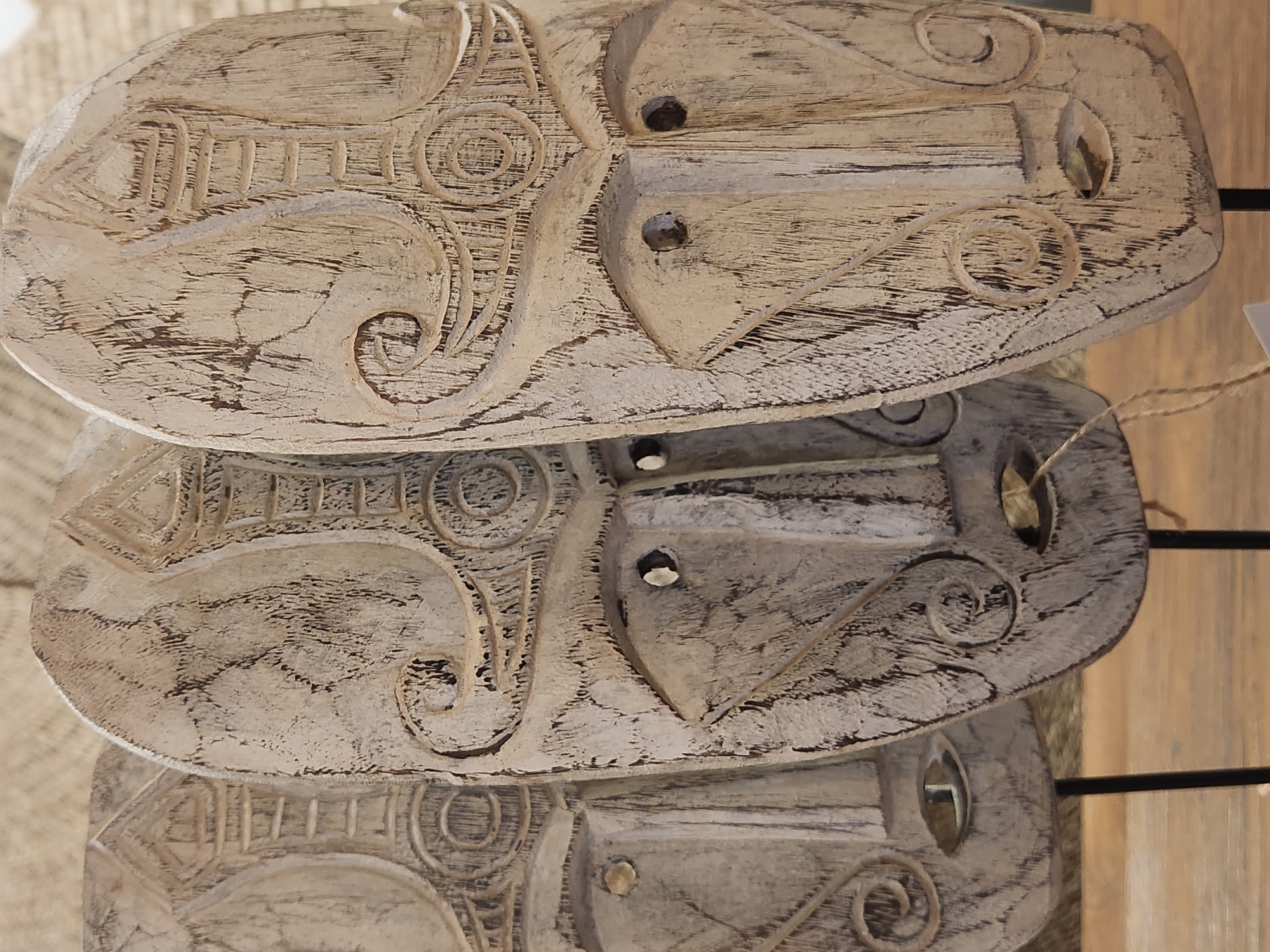 The image displays three carved wooden masks with an aged, distressed finish. The masks are positioned in a row, with the closest mask on the right and two others receding into the background. Each mask features intricate carvings, including patterned headdresses, stylized eyes, and swirling designs around the temples. The masks have visible eyeholes and mouth openings. They are displayed on black stands, and a piece of twine is visible hanging from the middle mask. The background appears to be a textured surface, possibly woven material. The lighting is soft, casting subtle shadows and highlighting the textures of the wood. There are no people present in the image. The masks appear to be decorative items, possibly for sale or display in a shop or market. The specific location in Maitland, United States of America, cannot be determined from the image alone.