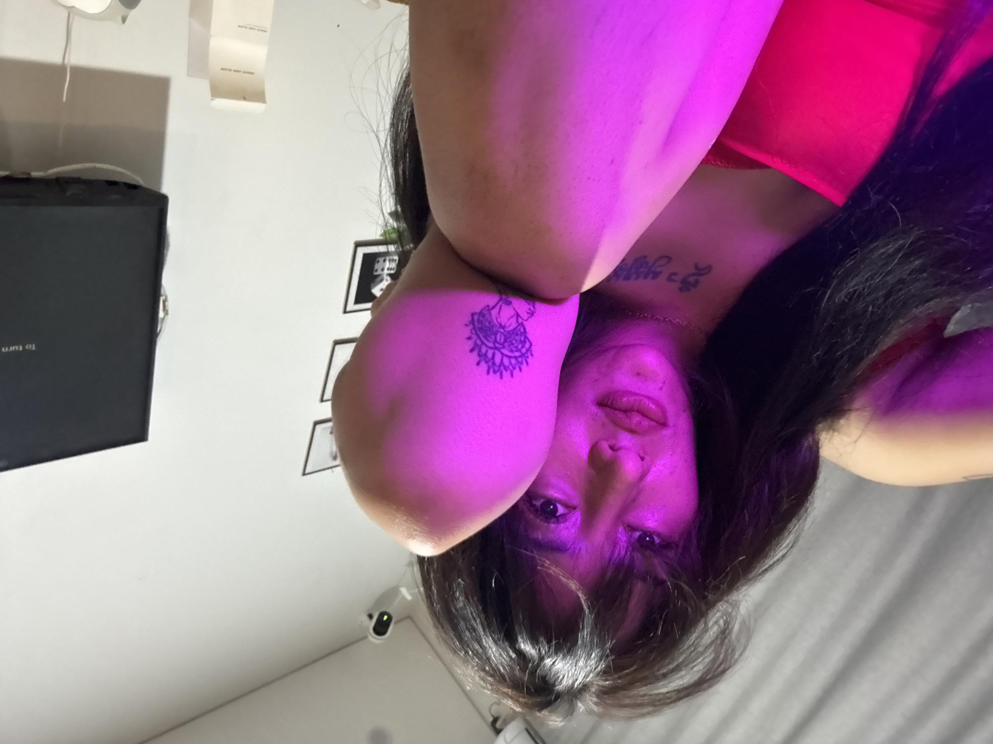 An overhead shot captures a person lying on their back, looking directly up at the camera. The scene is dominated by vibrant pink and purple lighting that illuminates the person's face and a raised arm. The person has dark hair that frames their face and spreads around their head. They wear a red garment, possibly a top or lingerie.
A prominent tattoo, depicting an intricate, stylized figure resembling an Indian deity or a mandala, is visible on their inner right forearm. Another tattoo, featuring text in an unidentifiable script, is visible on their upper chest, partially obscured by the garment. Their expression is direct and composed, with slightly parted lips.
The background reveals an indoor setting. To the person's left, a white wall holds several framed pictures and a black rectangular object, likely a monitor or TV, displaying the partial text "To turn...". A white paper tag hangs from the wall, clearly showing the words "SAUCE AND GLOW". A small white security camera or similar device is mounted on the wall in the lower left portion of the image. To the person's right, a gray fabric surface, possibly bedding, is visible. The distinct artificial lighting suggests an indoor setting, likely in the evening or a controlled environment.