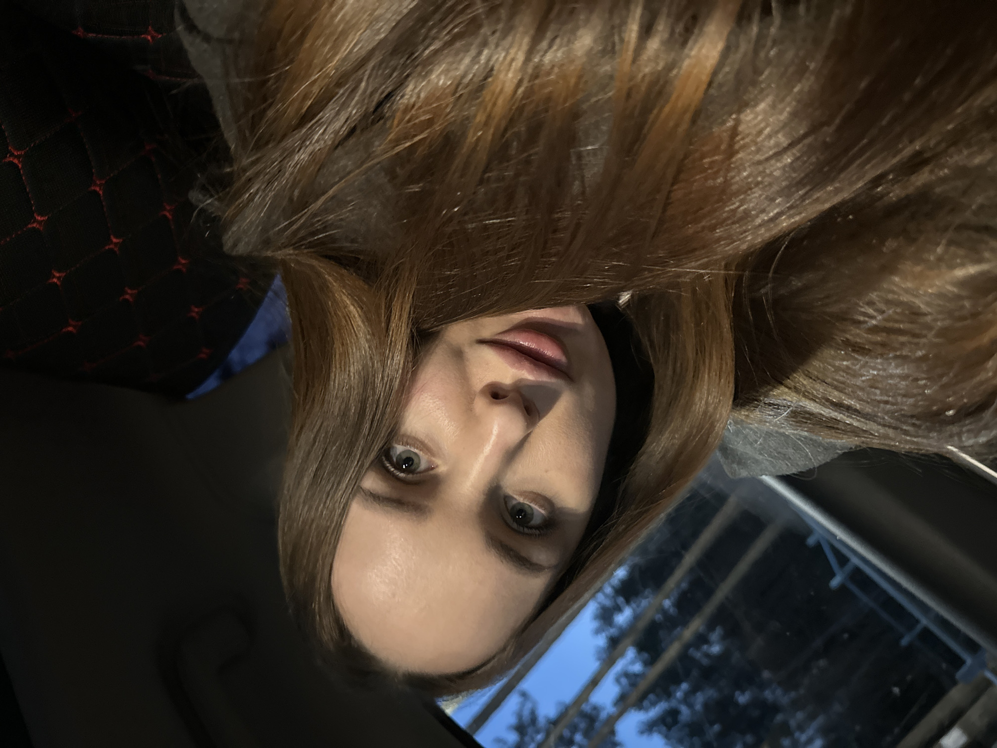 A woman with long, light brown hair is looking at the camera. She is in a car; the car seat cover is visible in the upper left. The window is on the right side of the image. The sky and trees are visible outside the window, suggesting an outdoor environment. The image does not contain sufficient information to ascertain the location in the city of Noviy, Russia.