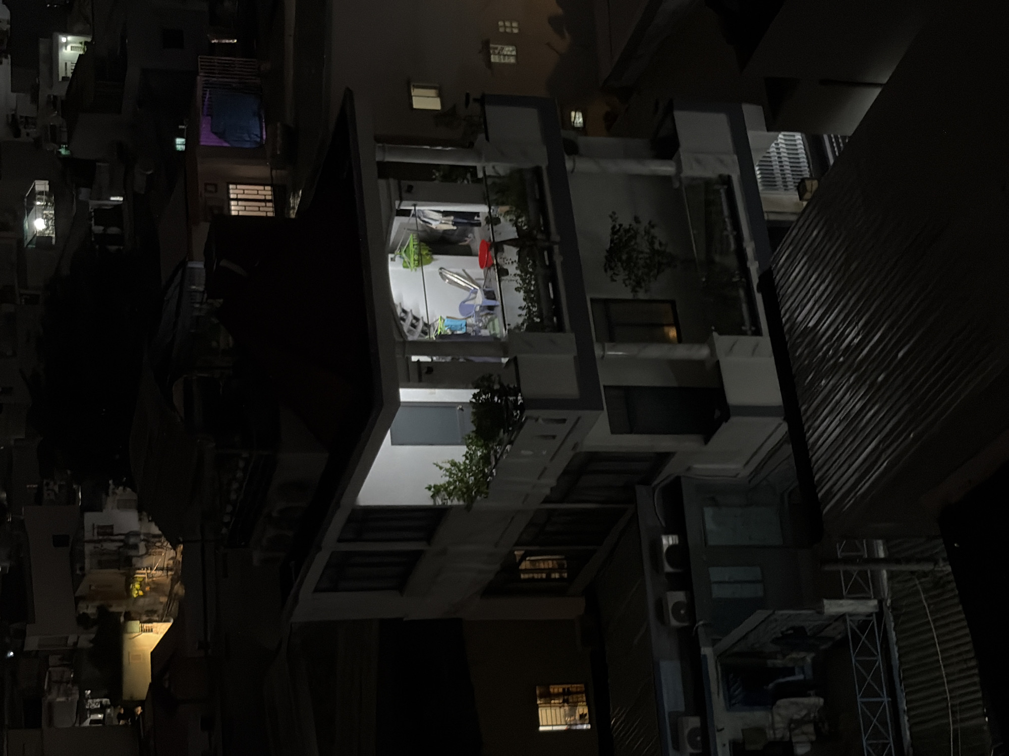 The image depicts a multi-story building at night in Thủ Đức, Vietnam. The building appears to be a residential or mixed-use structure, with balconies and windows visible. Some windows are illuminated, suggesting occupancy. On one balcony, clothes are seen hanging to dry, and there is a red basin and some chairs. The surrounding environment is dark, with other buildings and rooftops visible in the background, also partially lit by windows. The overall scene conveys a sense of urban nocturnal life.