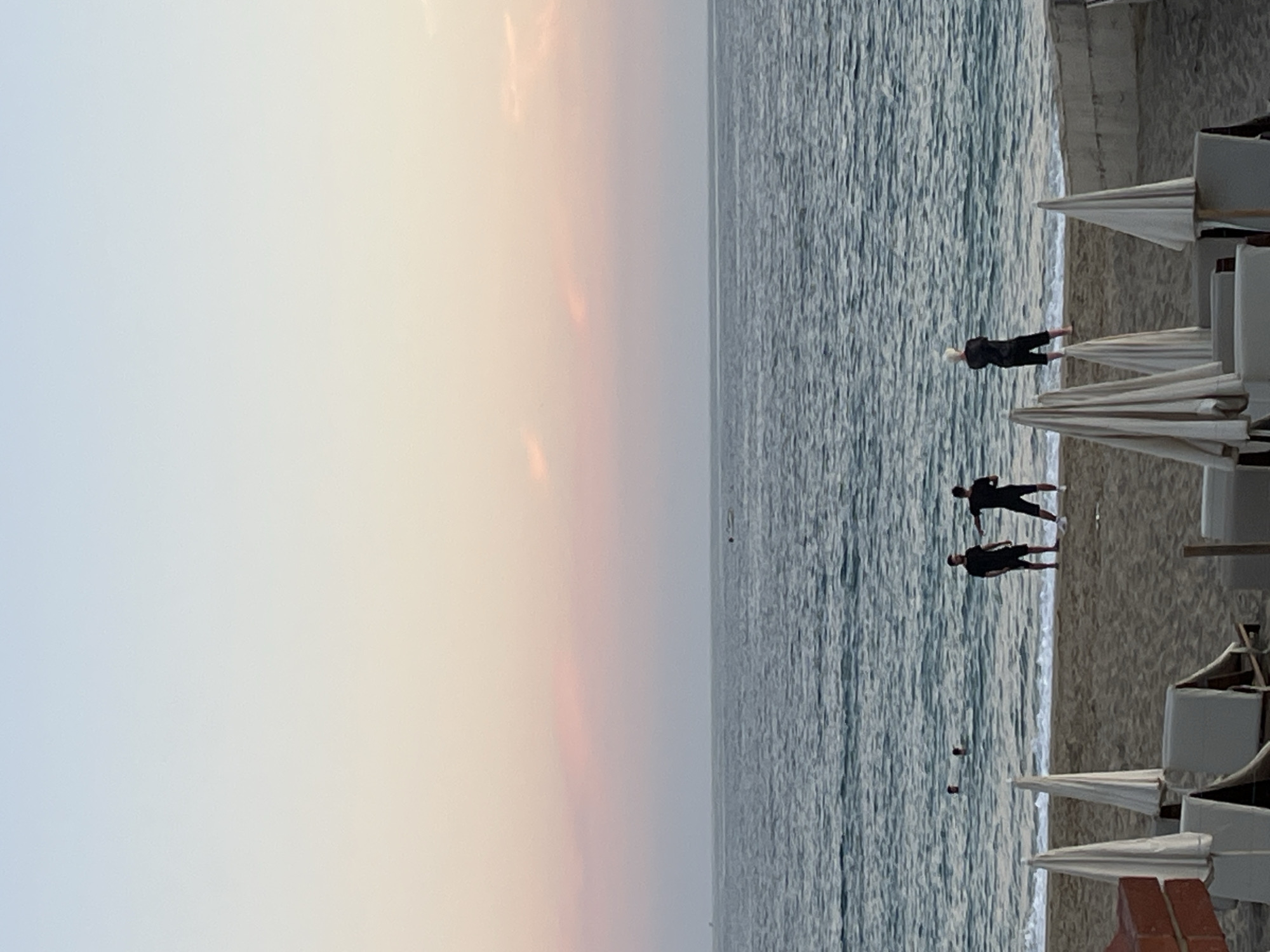 The image shows a beach scene in Alanya, Turkey at dusk. The sky exhibits a gradient of pale blue transitioning to shades of orange and pink near the horizon. The sea is textured with small waves. Three people are wading in the water near the shore. Along the beach, several closed beach umbrellas are lined up.