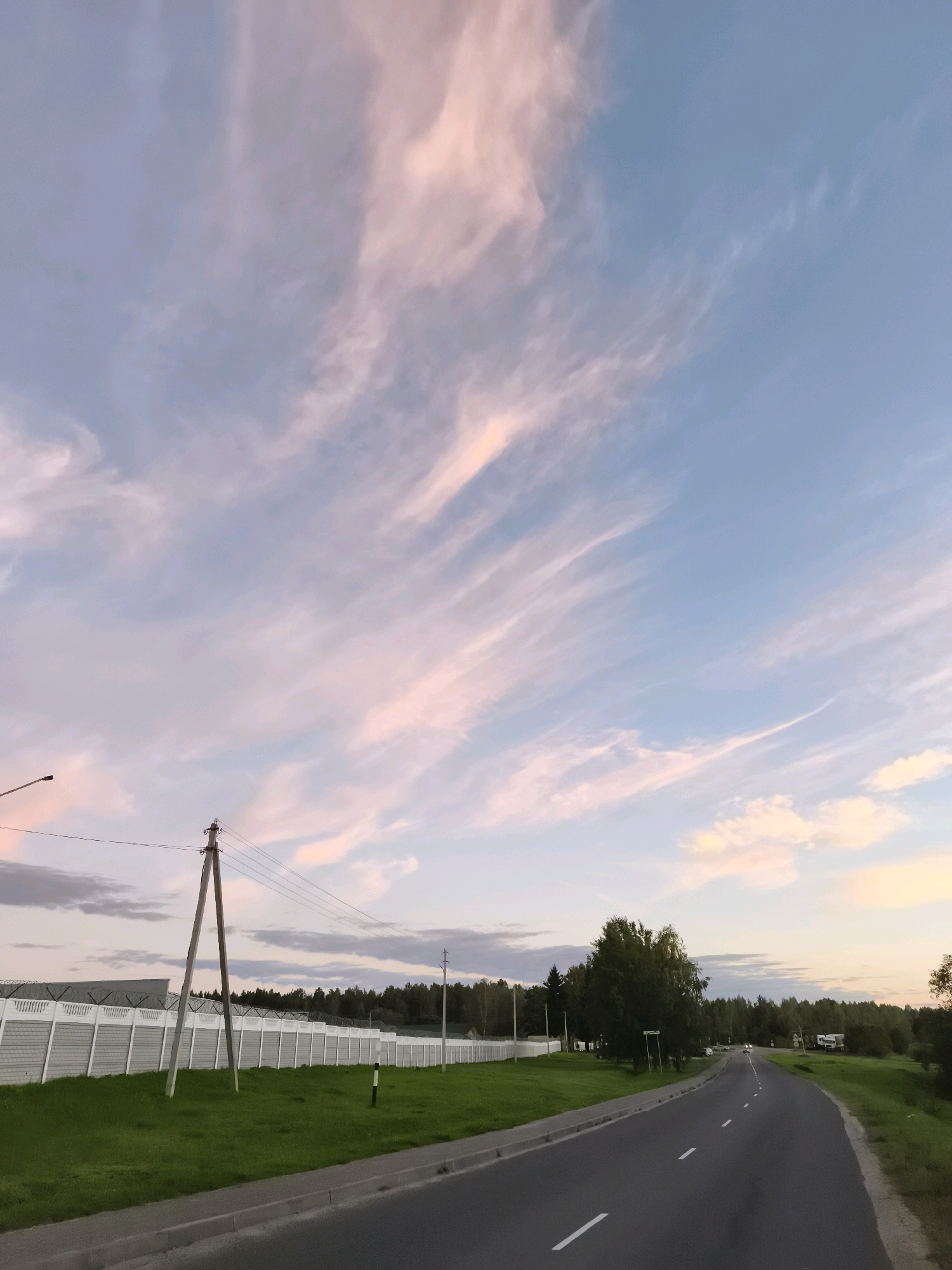 The image depicts a rural roadside scene in the vicinity of Горскі сельскі Савет, Belarus. A paved road with white dashed lane markers curves into the distance. On the left side of the road, a well-maintained grassy verge is bordered by a long, white concrete fence topped with barbed wire. A utility pole with wires stands prominently in the foreground, casting a shadow on the grass. Beyond the fence, a line of trees forms a dense forest, with a few buildings visible within the treeline. The sky above is a pale blue, transitioning to a soft pink and white hue due to wispy clouds, suggesting either sunrise or sunset. A few vehicles are visible in the distance on the road. The overall atmosphere is tranquil and serene.