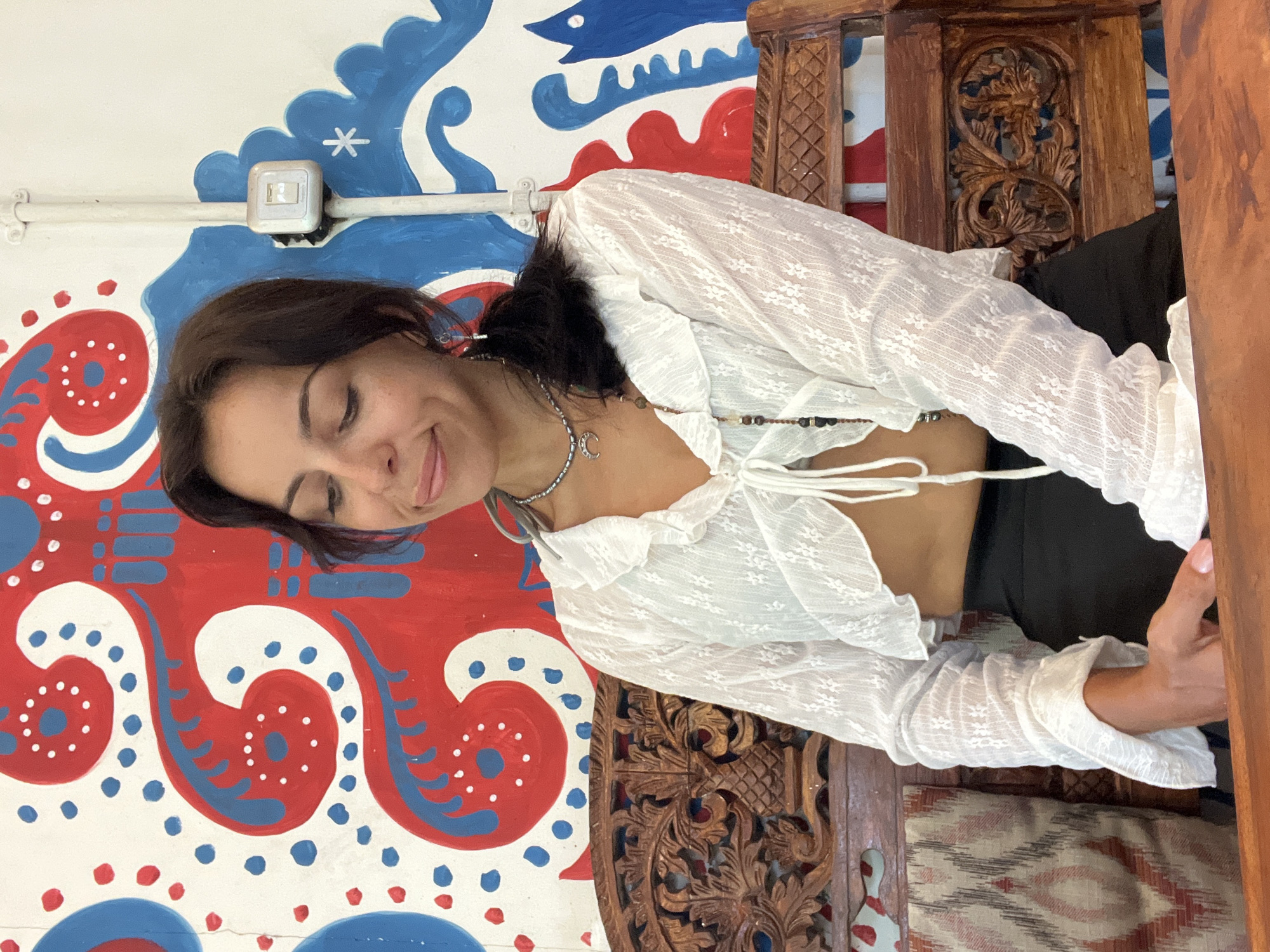A woman sits on a wooden chair in Kuta Selatan, Indonesia, against a wall painted with red and blue designs. She wears a white lace top, a silver necklace with a crescent moon charm, and dark trousers. Her eyes are closed, and she is smiling slightly. The chair has carved details. An electrical outlet is visible on the wall above the mural.