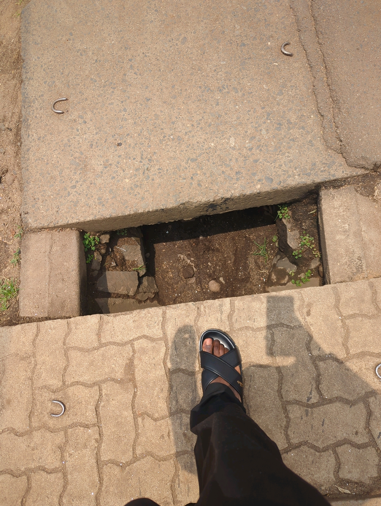 The image shows a close-up, high-angle view of a person's left foot wearing a black sandal, standing on a patterned concrete walkway in Musanze District, Rwanda. The foot is positioned next to an open concrete drainage channel. Within the channel, there is exposed soil, rocks, and small green plants. In the background, a large concrete slab is visible, with at least two U-shaped metal fixtures embedded in its surface. The concrete slab appears to be a manhole cover or similar infrastructure. The walkway is paved with interlocking concrete blocks. A shadow cast by the person and an unseen object is visible on the walkway. The scene suggests an outdoor urban environment, possibly a street or pathway, with evidence of civil infrastructure.