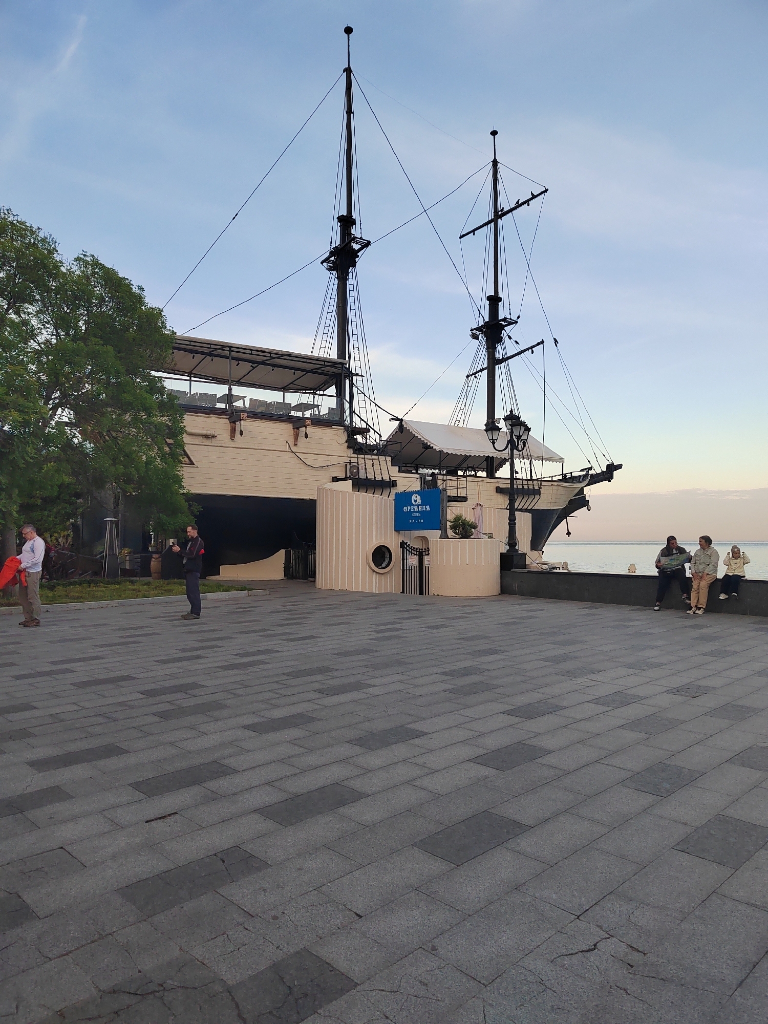 The image depicts a large, stationary ship-like structure situated in an urban environment. The structure resembles a vintage sailing vessel, complete with masts and rigging, but it is anchored to the ground and appears to serve as a building or establishment.
The scene is observed in the city of Unknown, Unknown, as evidenced by the characters on the blue sign that read in part "Ореанда Отель, HA-70."
The environment includes a paved plaza or promenade leading up to the structure, with several people present. On the left, two individuals are standing near a tree, one holding a red object. On the right, three people are seated on a low wall overlooking what appears to be the sea. The sky is clear with slight tonal color changes, indicating either sunrise or sunset.