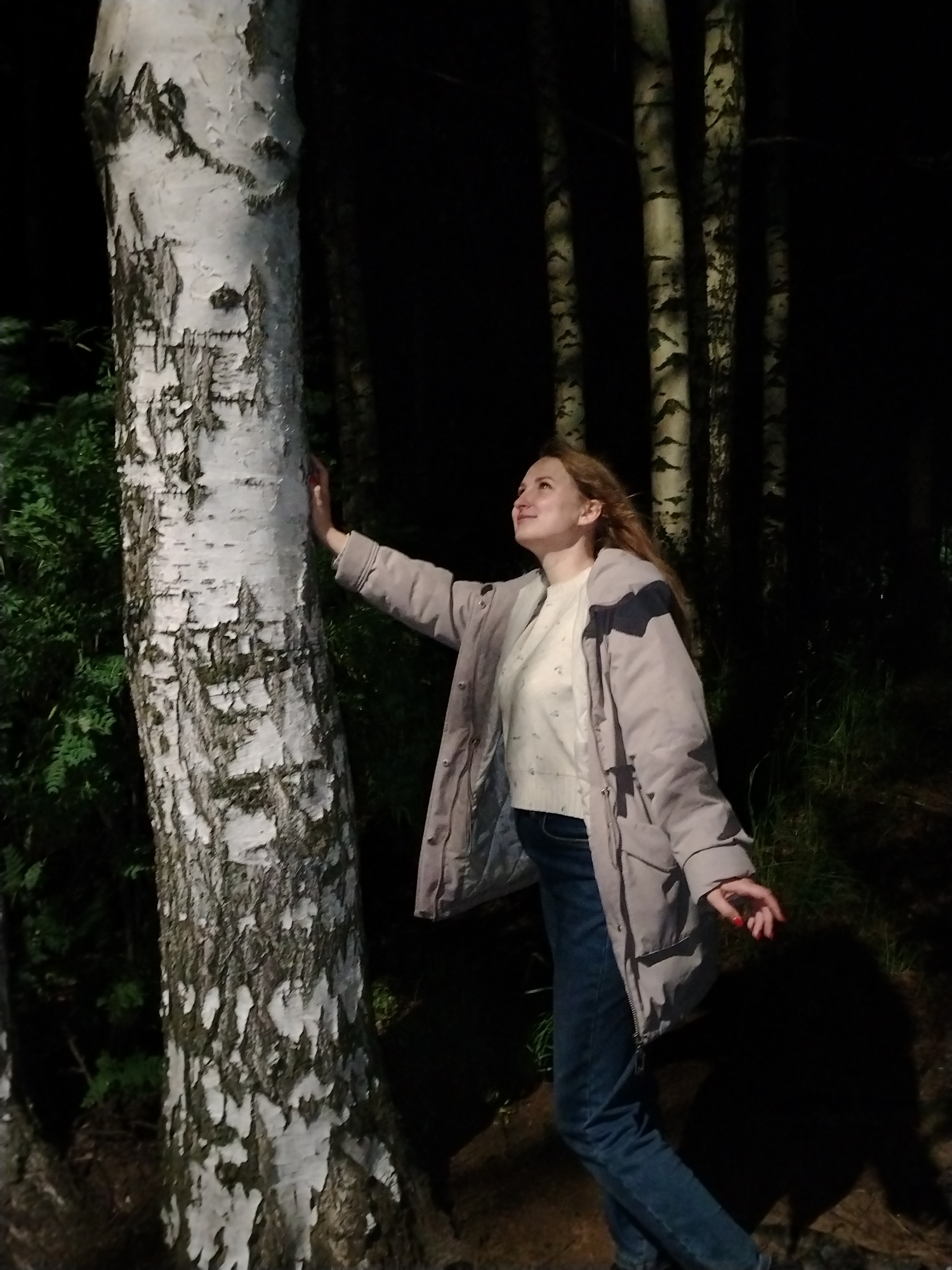A young adult female, with light-colored hair, stands outdoors at night in a wooded area within Chaikovsky, Russia. She is positioned slightly to the right of a prominent birch tree in the foreground, facing towards the right side of the frame with her head tilted upwards and a smiling expression. Her left hand is placed flat against the white, peeling bark of the birch tree trunk. She wears a light grey or beige padded jacket with darker grey or black accents on the shoulders and collar, a light-colored, possibly cream, long-sleeved top underneath, and blue jeans. Her fingernails on the visible right hand are painted red. The environment is dark, suggesting nighttime, with the immediate foreground and the subject illuminated by an artificial light source, possibly a flash. Behind the primary birch tree on the left, dark green foliage is visible. In the background to the right, several other birch trees with characteristic white bark and dark markings are discernible against the deep darkness. The ground appears uneven with some dark vegetation.