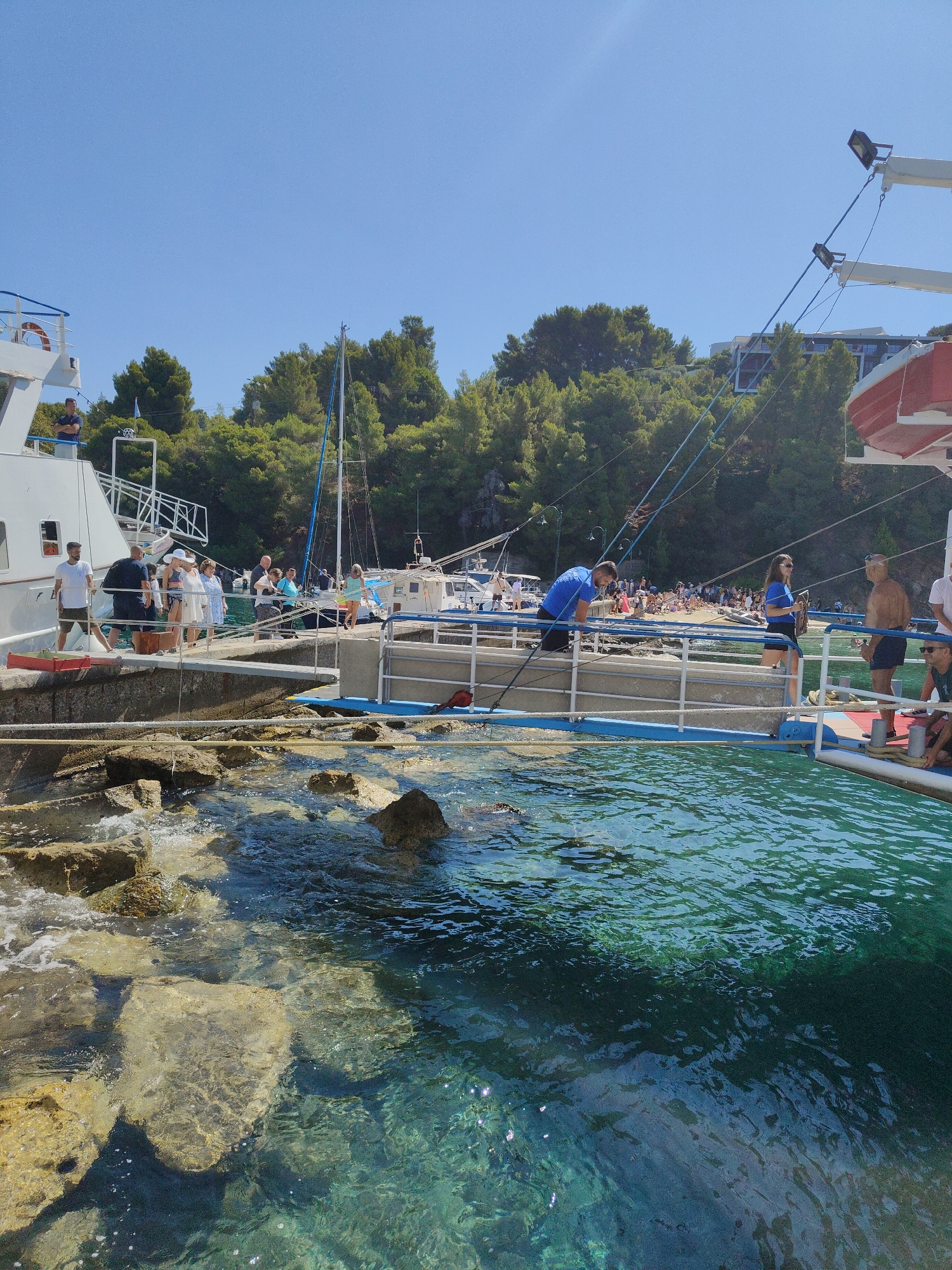 A group of people are disembarking from a white ferry onto a stone pier. The ferry has a red life raft attached to its side. The pier is lined with rocks and the water is clear and blue. In the background, trees and buildings are visible. The sky is clear and blue.