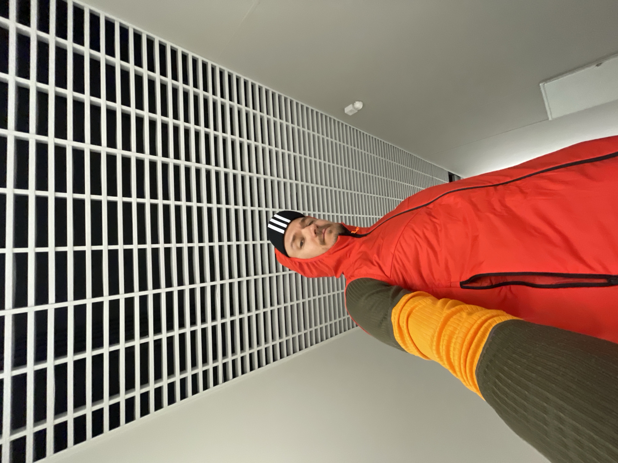 A man in a red jacket and a black and white striped beanie is standing indoors. He has on what appear to be yellow and green ribbed sleeves or arm warmers. The background consists of a white wall with a white grid-like pattern and a white ceiling. The location is not identifiable as Izhevsk, Russia from the image alone.