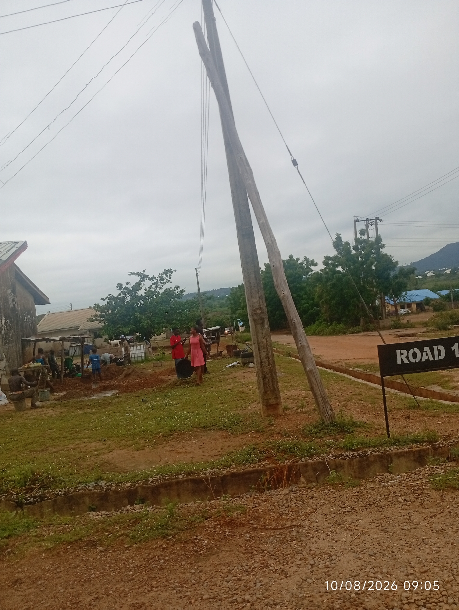 An outdoor, eye-level, medium shot captures a construction site in a rural or undeveloped area. The sky is overcast and a dull grey, suggesting a cloudy day. In the foreground, a gravelly path leads to a concrete drainage ditch. Two large wooden poles, leaning precariously, are prominent in the center and left of the frame, with electrical wires stretching across the sky.
The middle ground depicts a scene of activity. Several individuals are engaged in what appears to be construction work, with piles of red earth and construction materials visible. One person is near a water tank, and others are working with shovels and buckets. Two people, one in a red shirt and the other in a pink dress, stand in the middle of the scene, seemingly observing the work. In the background, there are several buildings with corrugated metal and blue roofs, surrounded by lush green trees. A white goat can be seen near one of the buildings. Further in the distance, a mountain range rises against the horizon.
To the right of the frame, a black sign with white lettering reads "ROAD 1". The bottom right corner of the image displays a timestamp: "10/08/2026 09:05". The overall impression is one of ongoing development in a semi-rural setting.