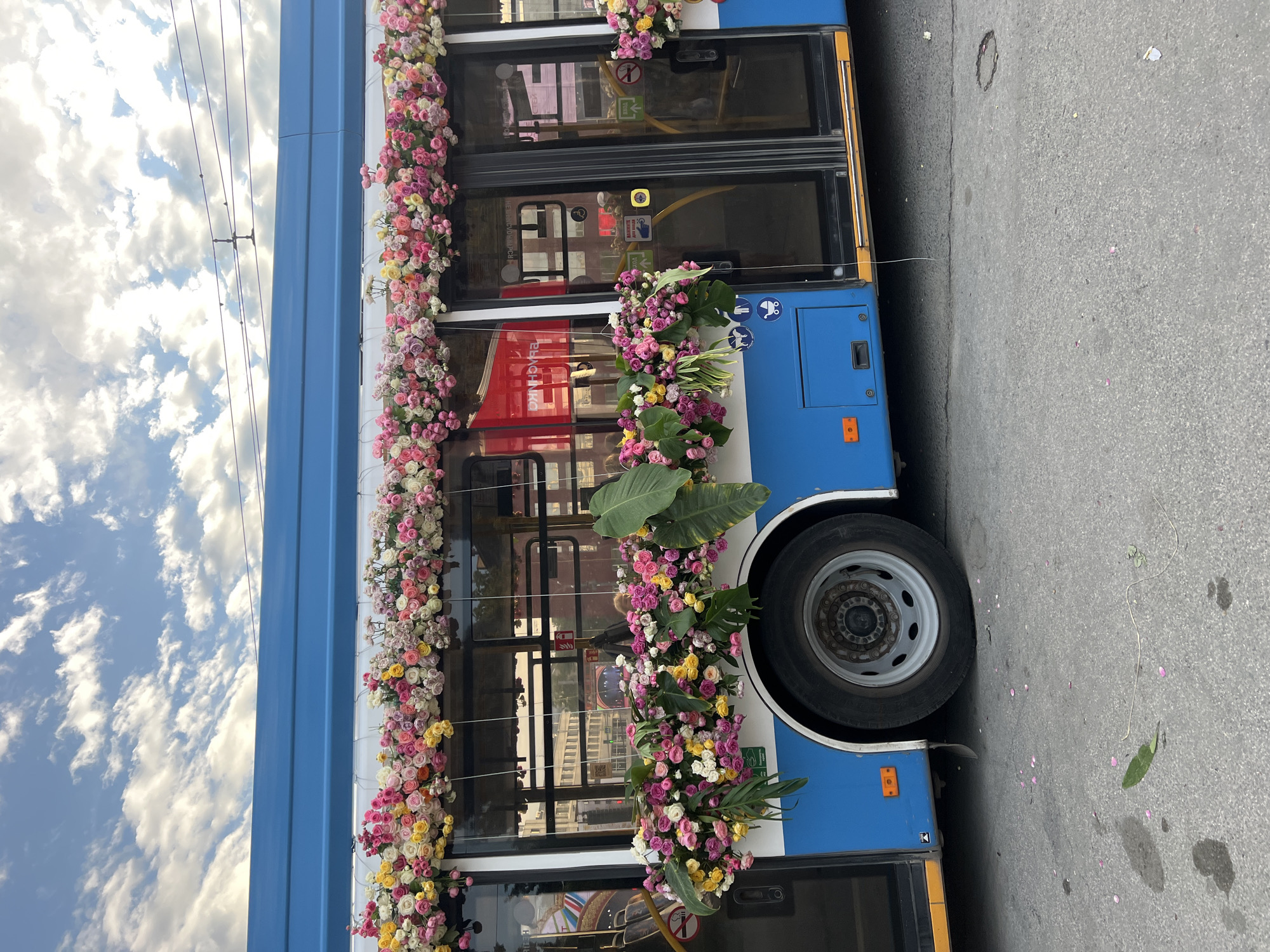 A blue and white bus is decorated with abundant floral arrangements along its top and sides. The flowers are predominantly pink, white, and yellow roses, with some greenery. The bus has tinted windows and double doors on the side. The scene appears to be in an urban environment, possibly a street or depot, with power lines visible in the sky. The ground is paved and shows some scattered flower petals and leaves. There are no people clearly visible in the image. The image does not provide any specific details that would allow for identification of the location in the city of Novosibirsk, Russia.