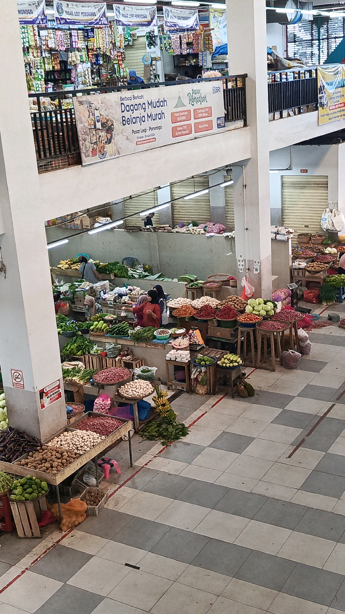 This image depicts an indoor market scene, likely Pasar Legi Ponorogo, in Ponorogo, Indonesia, as suggested by the banner in the upper portion of the image. The market is filled with stalls selling a variety of fresh produce.
In the foreground, to the left, a stall displays an assortment of vegetables, including dark purple eggplants, green cucumbers, and piles of red onions and white onions. Next to these are baskets of potatoes and an orange bag filled with produce. Further to the right, there are more stalls laden with various fruits and vegetables such as tomatoes, chili peppers, and leafy greens. Several people are visible browsing and interacting at these stalls.
The market environment is characterized by a tiled floor in a checkered pattern of gray and white. Elevated walkways and railings are present, suggesting a multi-level market structure. Banners with text, some in Indonesian, are hung throughout the market, including one prominently displaying "Pasar Legi - Ponorogo." Several stalls have wooden tables and baskets for displaying their goods. In the background, closed metallic shutters indicate shopfronts or storage areas. A woman wearing a hijab and an orange top is seen attending to customers at one of the produce stalls. Other individuals in the background are either shopping or working. The lighting appears to be a mix of overhead fluorescent lights and natural light filtering in from unseen openings.