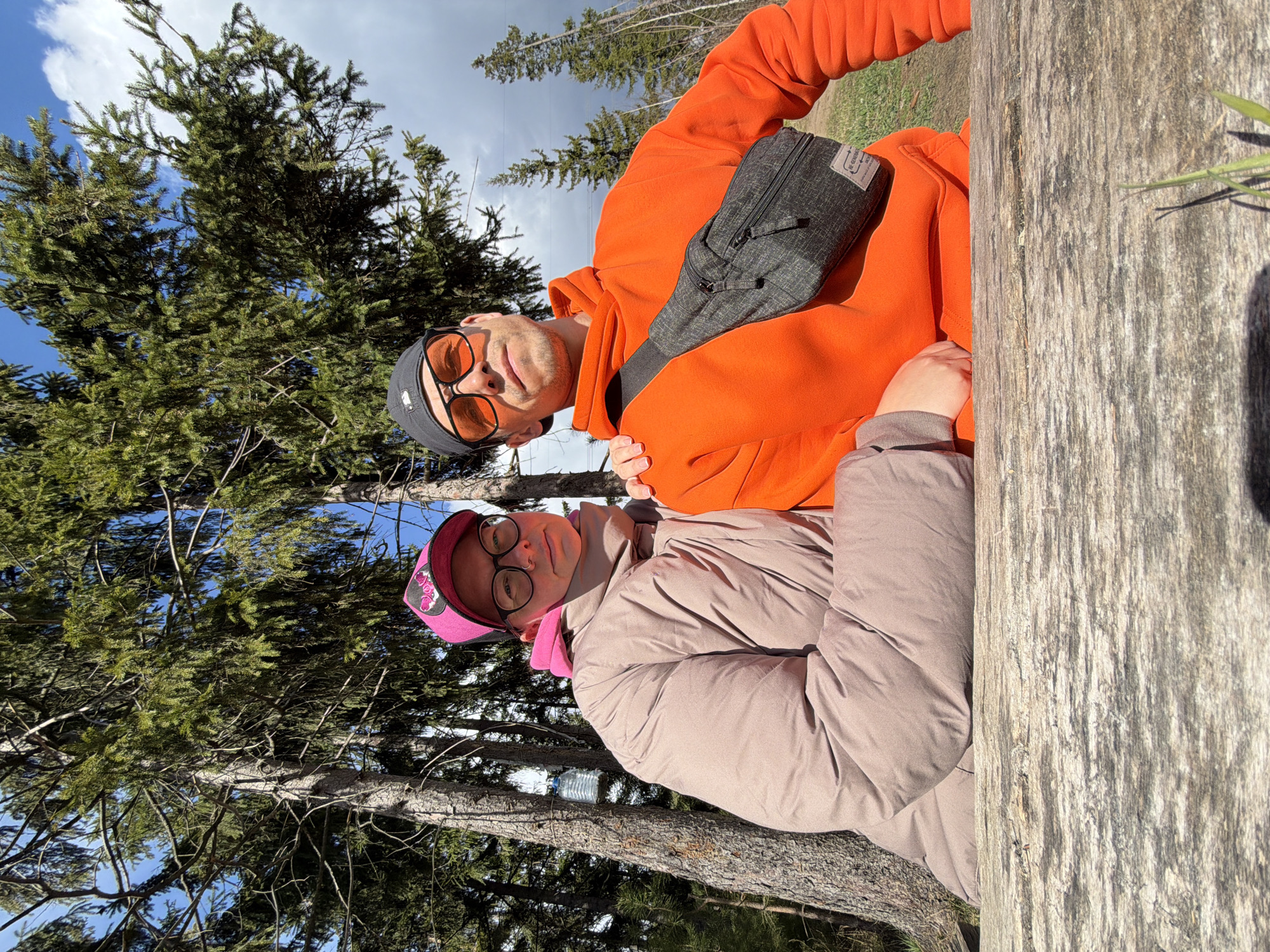 Two individuals are captured in an outdoor setting, likely a forest or wooded area, in Chaikovsky, Russia. The image is oriented sideways, with the subjects appearing to lean against a large, weathered wooden log that occupies the right side of the frame. Numerous coniferous trees with green needles are visible in the background against a bright sky with patches of white clouds and blue.
The person on the left is a male, wearing an orange hooded sweatshirt, a dark grey beanie, and orange-tinted sunglasses. A dark grey fanny pack or sling bag with a visible brand label "RE:SISTANT" is worn across his chest. He has light facial hair and looks directly at the camera with a neutral expression.
The person on the right is a female, dressed in a light brown or beige puffer jacket over a pink hoodie. She wears a pink baseball cap with a dark emblem, possibly a floral design, on the front, and prescription glasses. She also looks towards the camera with a neutral expression.
The bright daylight suggests it is daytime. Behind the female, a plastic water bottle is casually placed on a tree branch or log, indicating a relaxed outdoor activity. The overall scene conveys a casual outing in a natural environment.