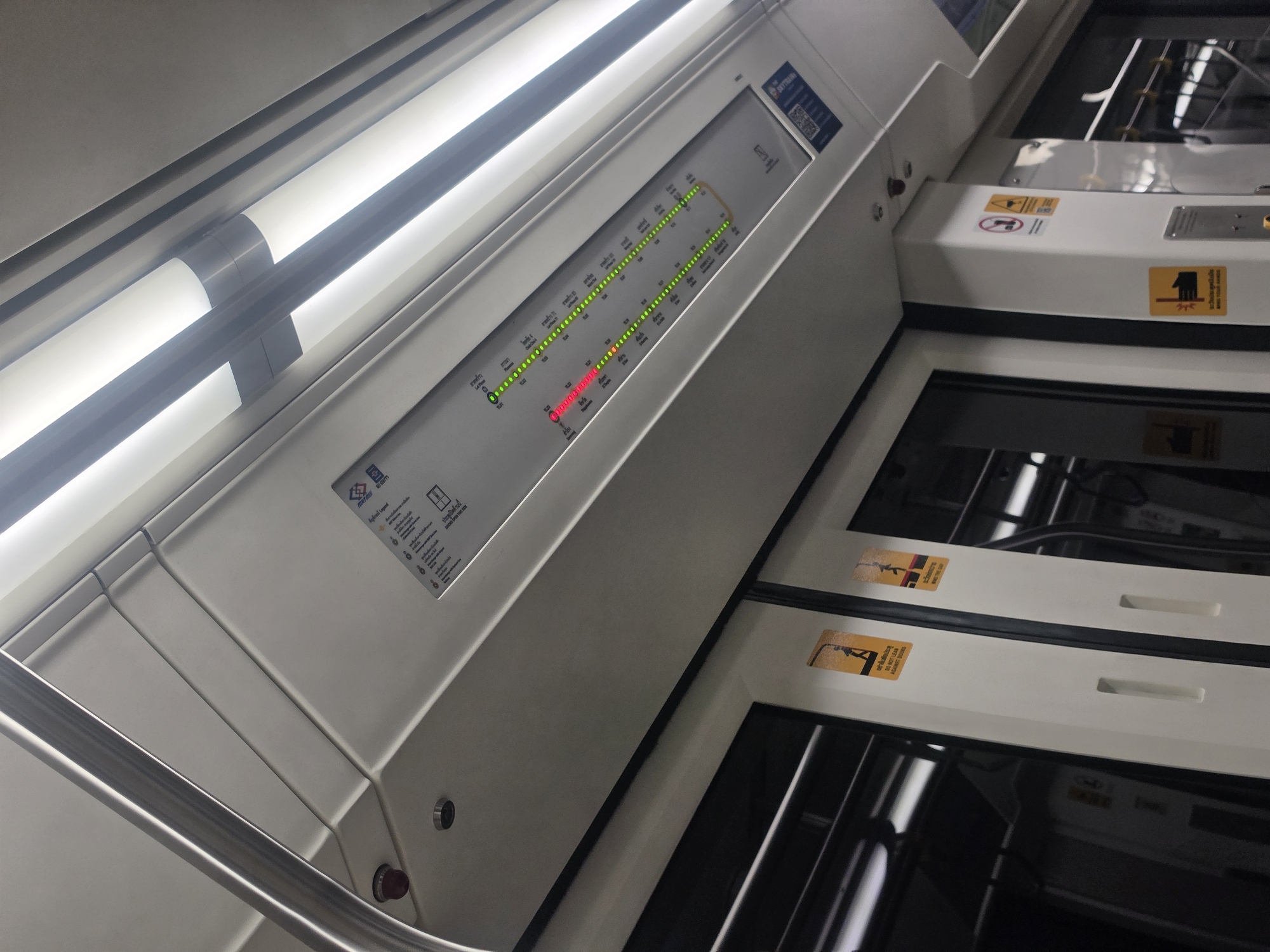 Inside a train car in Bang Mueang Subdistrict Municipality, Thailand, a subway map with LED lights indicates the train's current location. The map shows a series of stations, with green lights marking the stops already passed and a red light indicating the current station. Adjacent to the map are the train doors, featuring caution stickers. Overhead lighting is provided by fluorescent bulbs. A safety handrail is visible in the foreground.