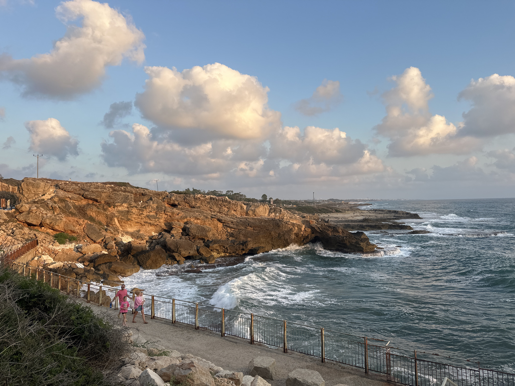 The image depicts a scenic view of the coastline in the Mate Asher Regional Council, Israel. The foreground features a paved walkway with a metal railing, bordered by rocky terrain and dense green bushes on the left. Three people, two adults and a child, are walking on the walkway. The two adults appear to be a couple, with one wearing a pink t-shirt and shorts, and the other a pink t-shirt and denim shorts. The child is wearing a pink t-shirt and shorts.
The walkway leads towards a rugged, orange-hued cliff face that dominates the left and central portions of the image. Waves crash against the base of the cliff, creating white foam that contrasts with the deep blue of the sea. The sea extends to the right, with more rocky outcrops and waves visible further along the coast. In the distance, the coastline continues, with some buildings and vegetation visible. The sky above is a clear blue, with scattered, fluffy white clouds, some of which are illuminated by the sun. The overall impression is a beautiful coastal landscape under a bright sky.