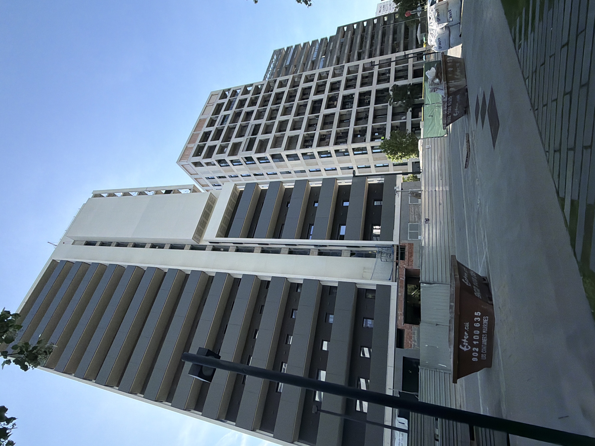 The image depicts a street-level view of modern buildings in l'Hospitalet de Llobregat, Spain, on a clear day with a bright blue sky. Two tall buildings dominate the frame. The first building has a white facade with dark gray vertical lines creating a pattern, and windows are visible between the lines. A second, more distant building with a grid-like design is visible above the first. In the foreground is a street or sidewalk, with a long dark gray container or dumpster visible. This container has the name "Ester.cat" and the phone number "902 100 635" printed on it, followed by the words "LDS CONTAINERS MARRONES". A tall, dark post or pole is also present in the foreground, stretching vertically. White sacks, possibly construction materials, are visible on the ground in the distance.
