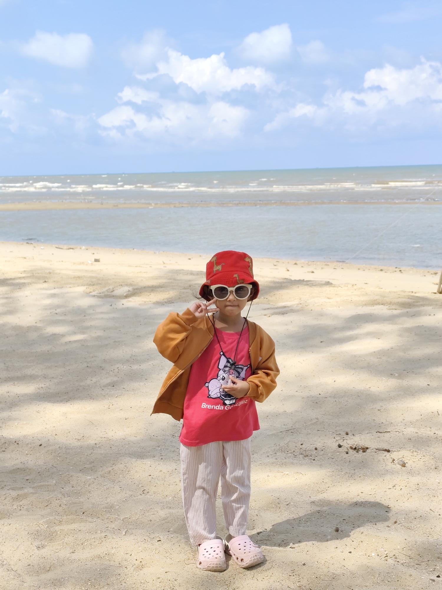 A young girl stands on a sandy beach, wearing a red bucket hat, sunglasses, a red t-shirt with a cartoon design, tan pants, and pink Crocs. She is facing the camera and holding her sunglasses with one hand. The ocean and sky with clouds are visible in the background. The image appears to be taken outdoors in natural light.